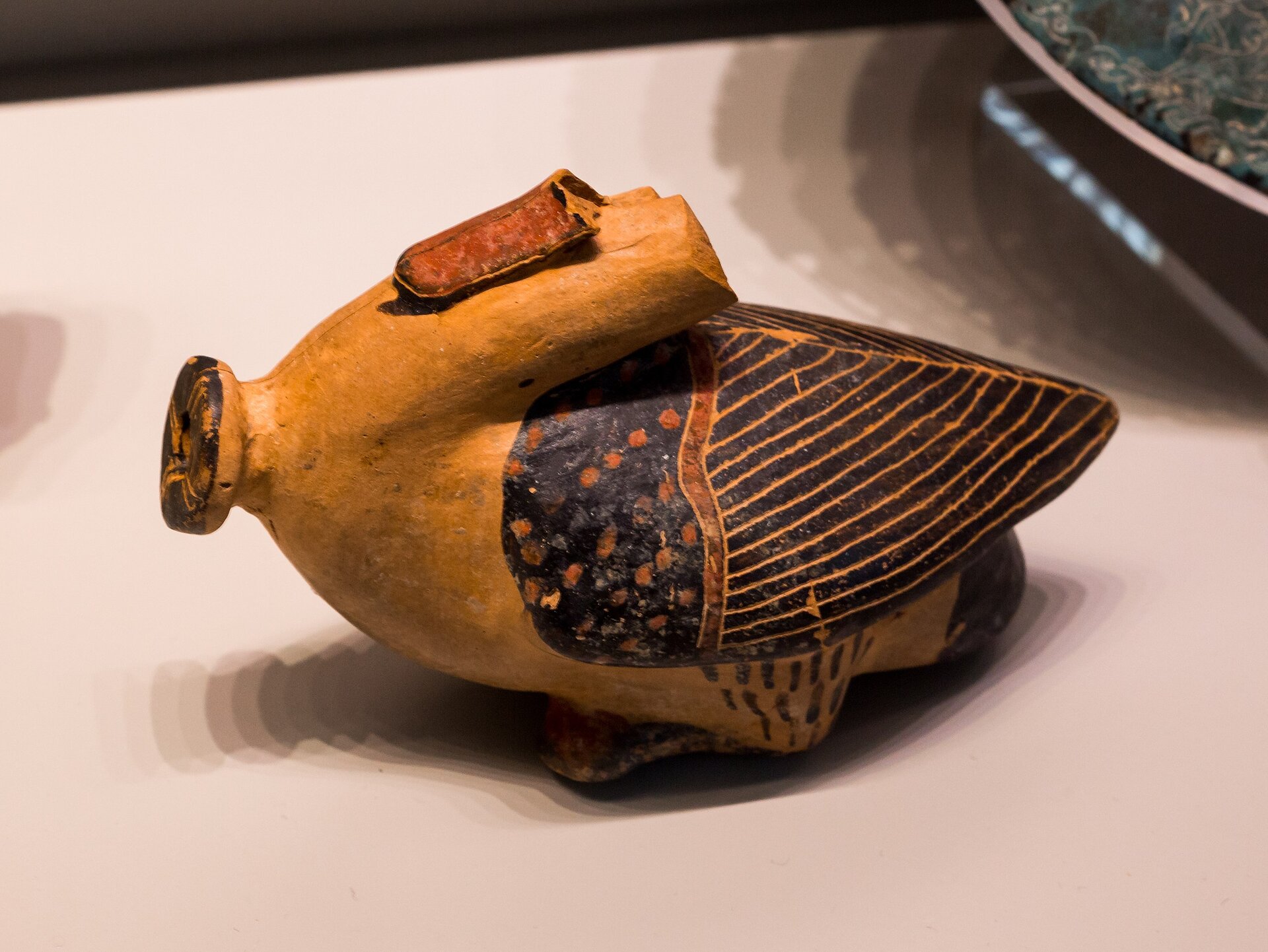 Fotografia autorstwa ArchaiOptix przedstawia starożytny aryballos - naczynie swoim kształtem przypominające gęś lub kaczkę bez głowy (na jej miejscu jest krótka szyjka do nalewania płynu). Aryballos został uformowany na kształt zwierzęcia, ma zarysowane kolorowe skrzydła, szyję oraz kuper. Fotografia została wykonana na jasnym stole.