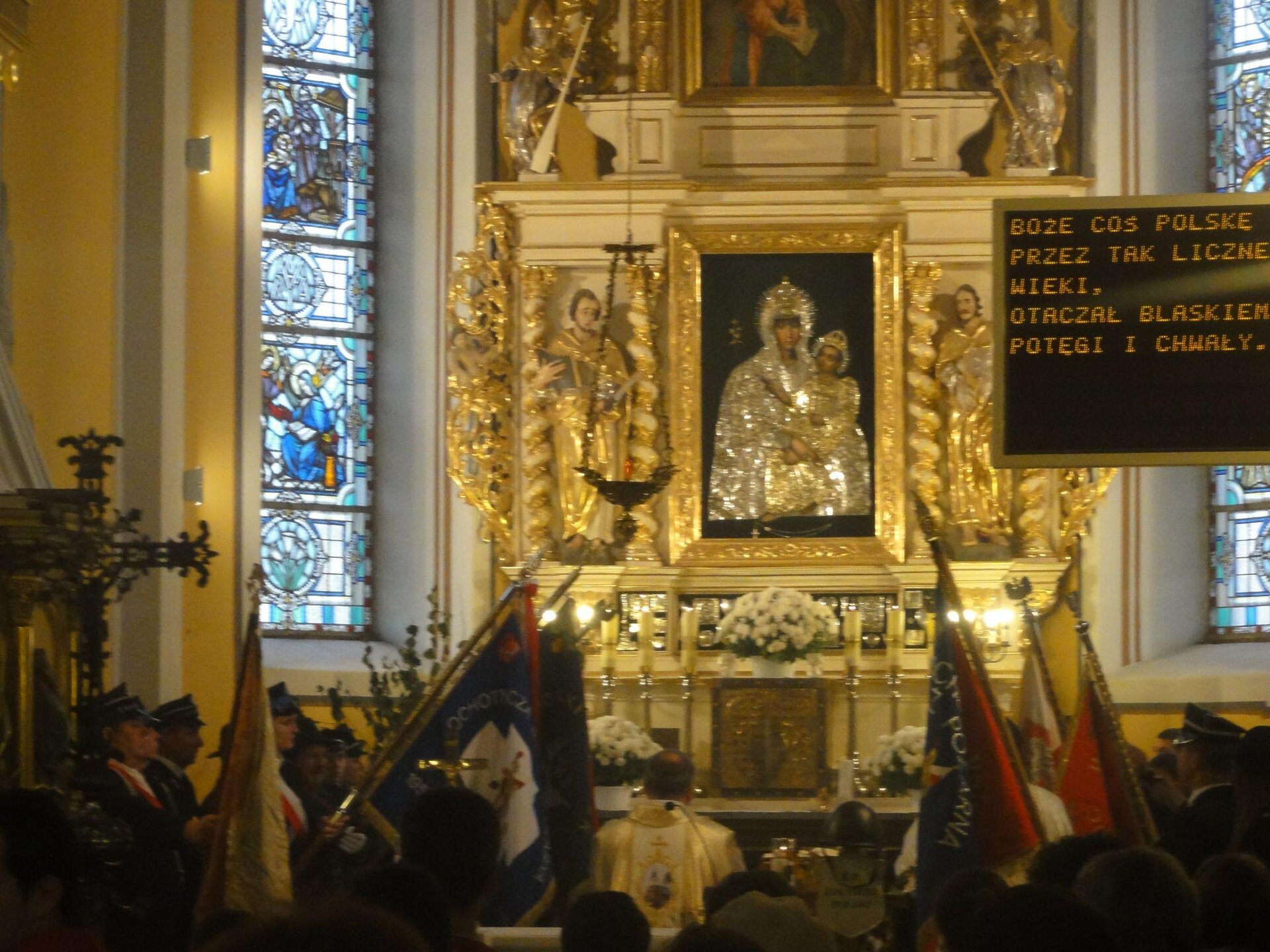 Zdjęcie przedstawia ołtarz główny. W jego centralnej części znajduje się obraz Matki Boskiej z Jezusem w złotej ramie. Po obu stronach obrazu znajdują się sylwetki mężczyzn ubranych w złote szaty. Nad obrazem znajduje się kolejny, również w złotej ramie, oraz sylwetki aniołów w złocie i srebrze. Po obu stronach ołtarza znajdują się podłużne okna, w których widoczne są witraże. Przed ołtarzem stoi ksiądz w złotej szacie, a także mężczyźni w mundurach z czerwono‑białą szarfą na piersi. Część z nich trzyma flagi, które pochyla w kierunku ołtarza. Z prawej strony zdjęcia widoczny jest telebim, na którym znajduje się pomarańczowy napis zapisany drukowanymi literami: "Boże coś Polskę przez tak liczne wieki, otaczał blaskiem potęgi i chwały".