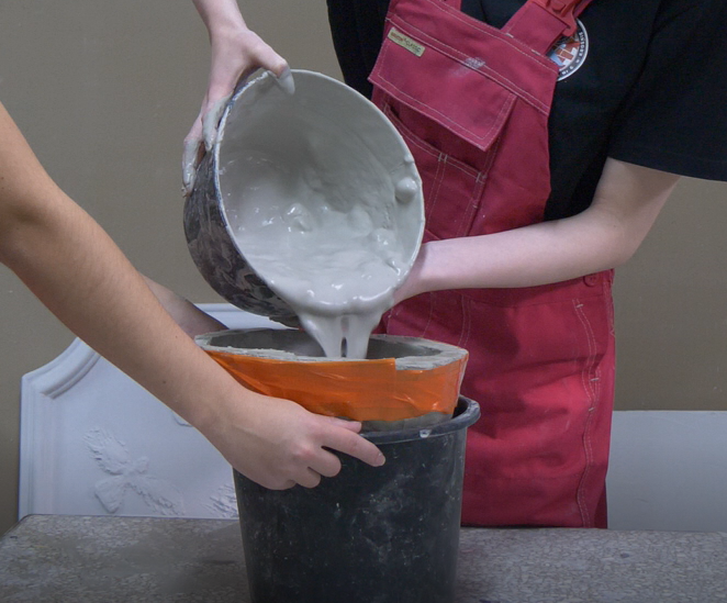 Zdjęcie przedstawia model detalu architektonicznego wykonany z gipsu. Jeden z pracowników wlewa z wiaderka gips do formy włożonej do wiadra. Drugi pracownik natomiast przytrzymuje wiadro by ustabilizować formę.