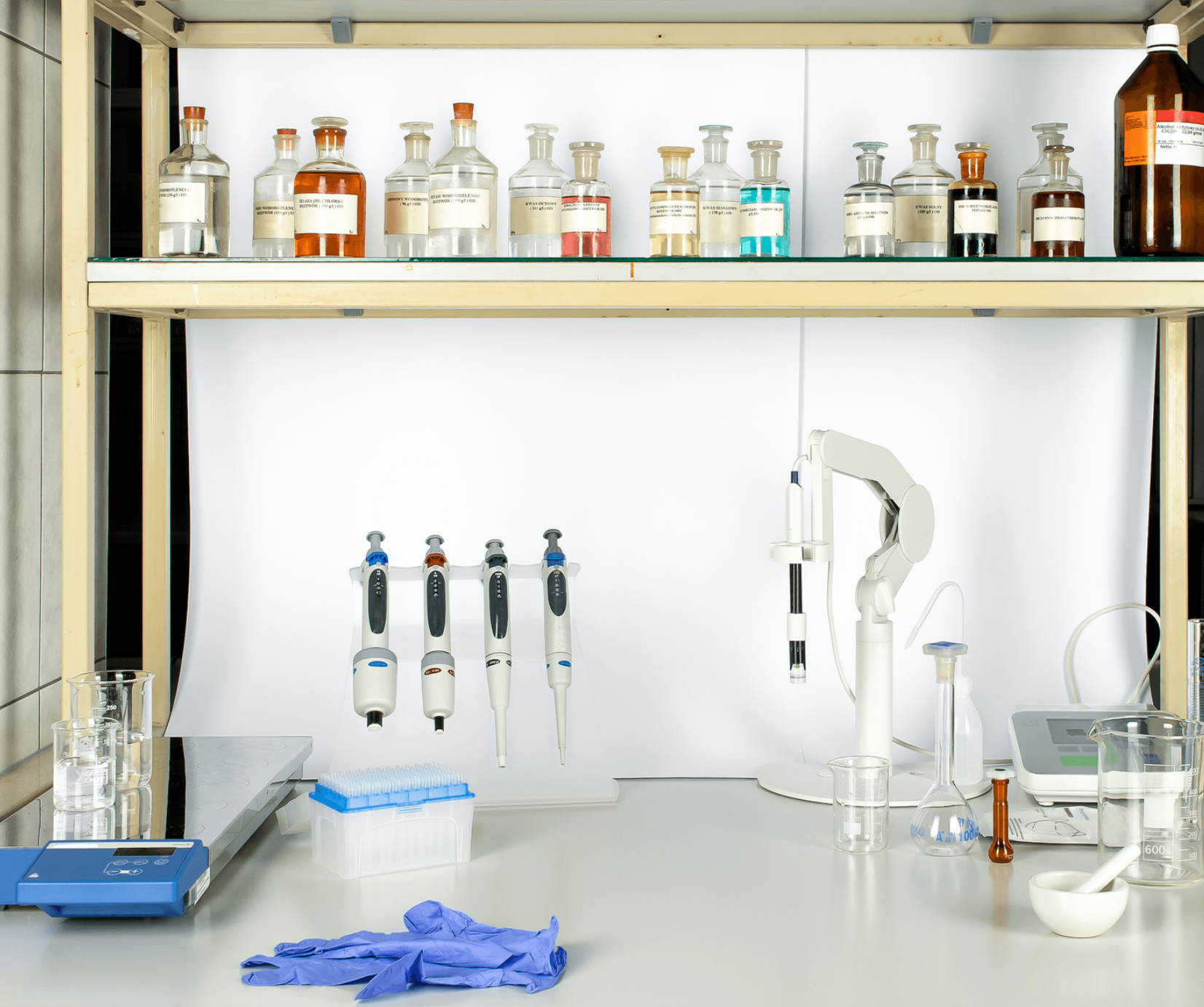 Zdjęcie przedstawia stanowisko do wykonywania reakcji chemicznych. Na górze znajduje się półka wypełniona odczynnikami chemicznymi w szklanych pojemnikach. Na stole widoczna jest aparatura: waga elektroniczna, stojak z pipetami automatycznymi, pehametr, szkło laboratoryjne oraz moździerz. 