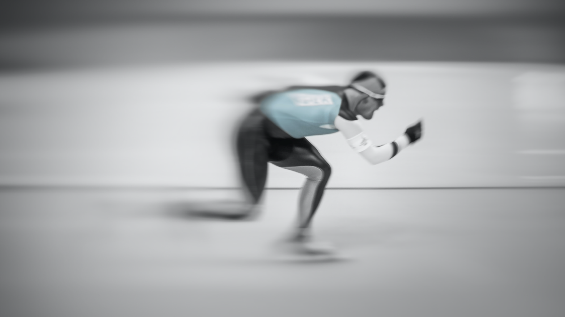 Zdjęcie przedstawia łyżwiarza szybkiego podczas jazdy po torze. Łyżwiarz ubrany jest w kombinezon i okulary. Zdjęcie jest rozmyte.