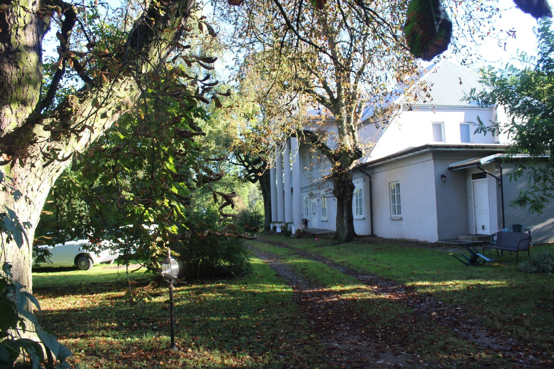 Zdjęcie przedstawia biały dworek ujęty od boku. Przed wejściem do dworku znajdują się białe kolumny. Dworek jest piętrowy, a jego dach jest spadzisty. Wokół dworku rośnie zielona trawa, a także liczne drzewa, które porastają zielone liście. Zdjęcie zostało wykonane w świetle dziennym.