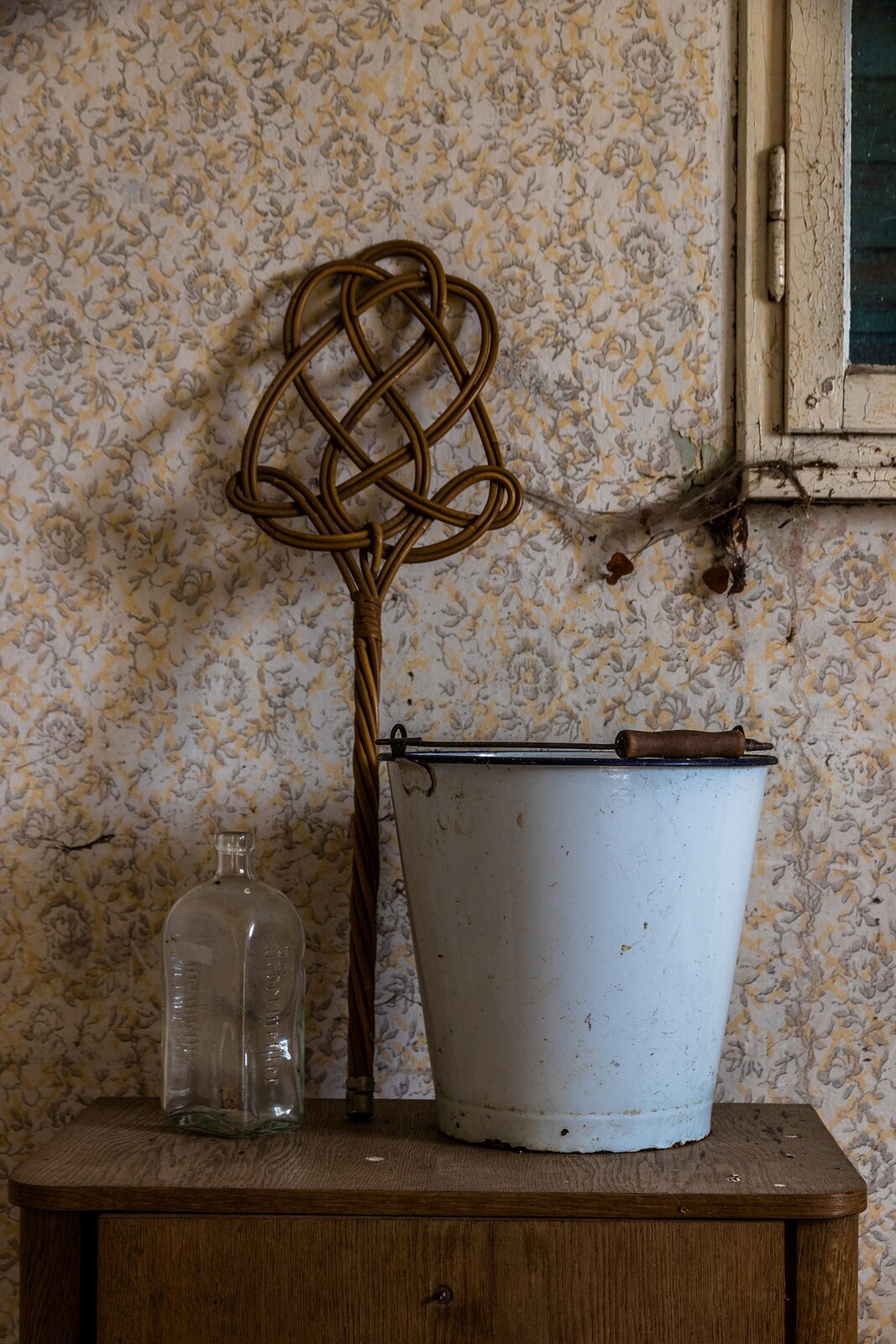 Zdjęcie przedstawia szklaną butelkę, wiklinową trzepaczkę i blaszane stare wiadro. Przedmioty te znajdują się na drewnianym stoliku na tle wzorzystej ściany z pajęczynami.