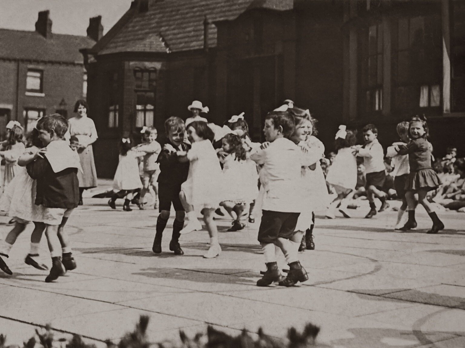 Czarno‑białe zdjęcie przedstawia dzieci w wieku przedszkolnym, które tańczą w parach na boisku/dziedzińcu. Dziewczynki ubrane są w jasne sukienki, a część z nich ma kokardę na głowie. Chłopcy są ubrani w ciemne, krótkie spodnie i mają albo jasne, albo ciemne marynarki z kołnierzami. W tle po lewej stronie widoczna jest stojąca kobieta, ubrana w bluzkę i spódnicę do łydek. Za nią i po prawej stronie widoczny jest duży, drewniany budynek ze spadzistym dachem krytym dachówką.