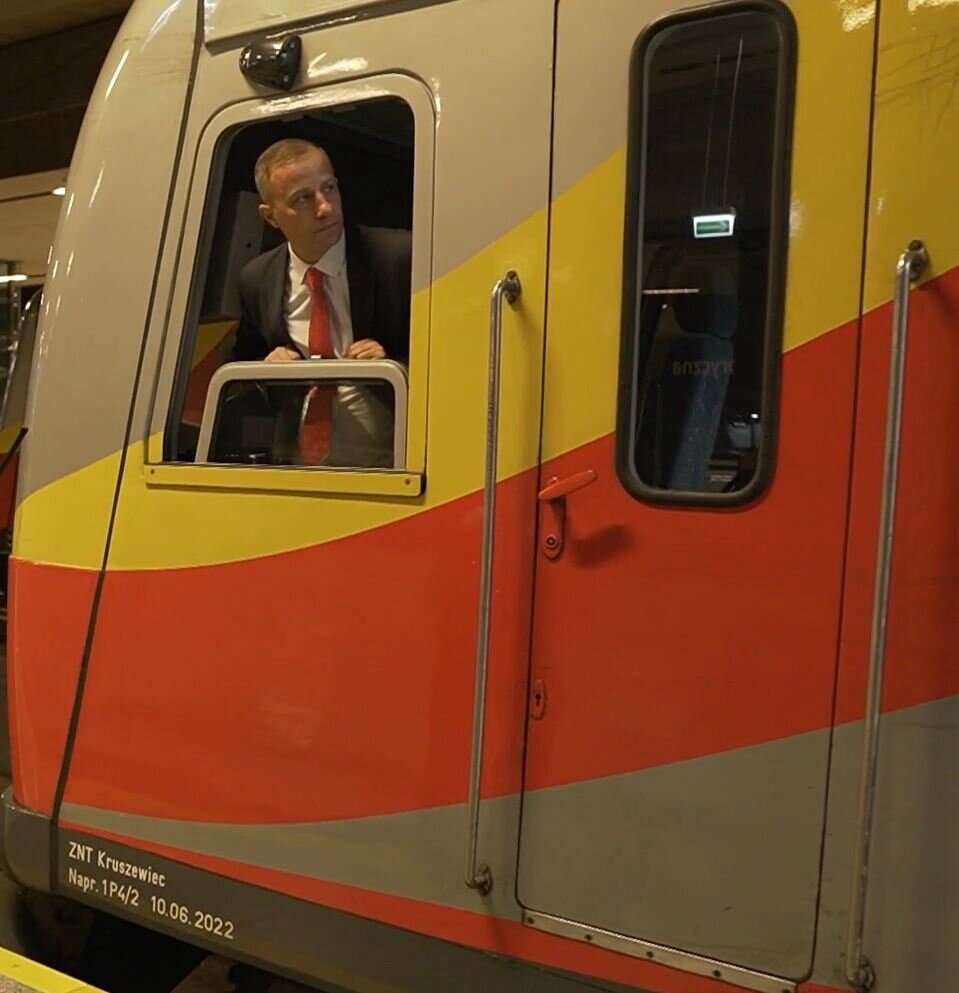 Zdjęcie przedstawia maszynistę. Mężczyzna w białej koszuli, czarnej marynarce i z czerwonym krawatem wygląda przez okno lokomotywy pociągu stojącego na dworcu.