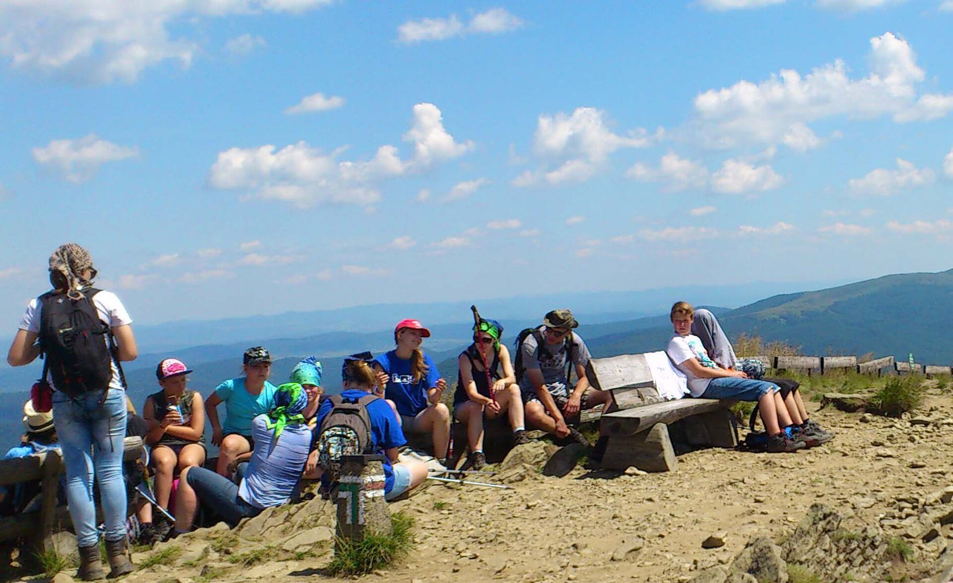Na zdjęciu grupa dzieci na górskim szlaku wraz z dwójką dorosłych opiekunów. Siedzą na ławkach lub na ziemi, kolorowo ubrani, większość z nich ma czapki na głowach chroniące przed intensywnym letnim słońcem. W tle rozległe tereny górskie z licznymi wzniesieniami i dolinami.