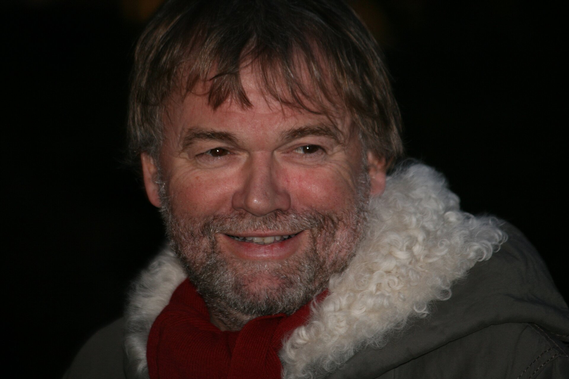 Kliknij, aby powiększyć. Zdjęcie przedstawia mężczyznę ujętego od ramion w górę. Ma dłuższe, brązowe włosy, które częściowo zakrywają mu uszy i czoło. Ma gęste, brązowe brwi, ciemne oczy i duży nos. Uśmiecha się lekko i odsłania szereg prostych, białych zębów. Na policzkach i brodzie ma krótki czarno‑biały zarost. Mężczyzna spogląda w prawą stronę. Ubrany jest w ciemnozieloną kurtkę, która ma biały kożuch wszyty w kapturze, a na szyi bordową chustkę. Tło za nim jest czarne.