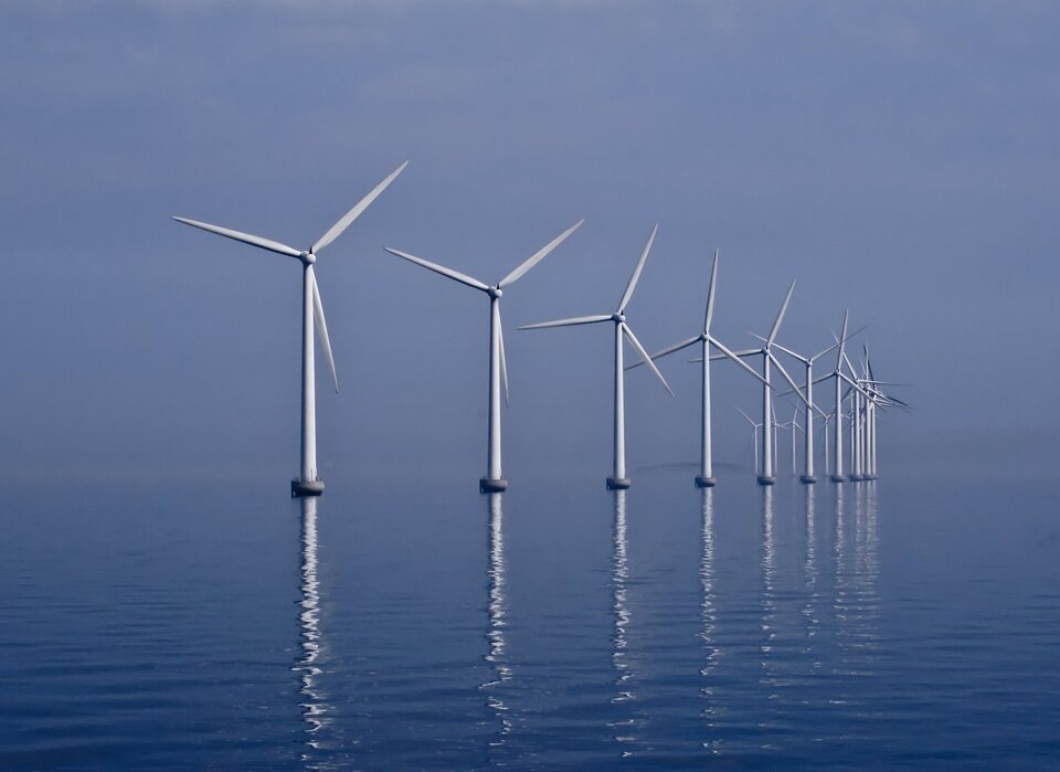 Zdjęcie przedstawia elektrownię wiatrową zlokalizowaną na wodzie. Tworzy ją kilkanaście trójramiennych wiatraków ustawionych na grubych słupach.