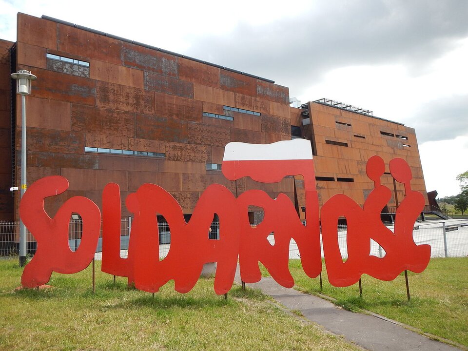Zdjęcie przedstawia duży napis Solidarność zapisany czcionką solidarycą. Napis stoi przed dużym budynkiem.