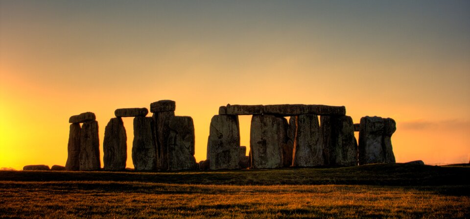 Zdjęcie, przedstawiające ustawione w okrąg kamienie megalityczne - pionowo, jak i poprzecznie ułożone na dwóch pionowo stojących kamieniach, ukazane na tle nieba ze światłem zachodzącego słońca. Zdjęcie współczesne. 