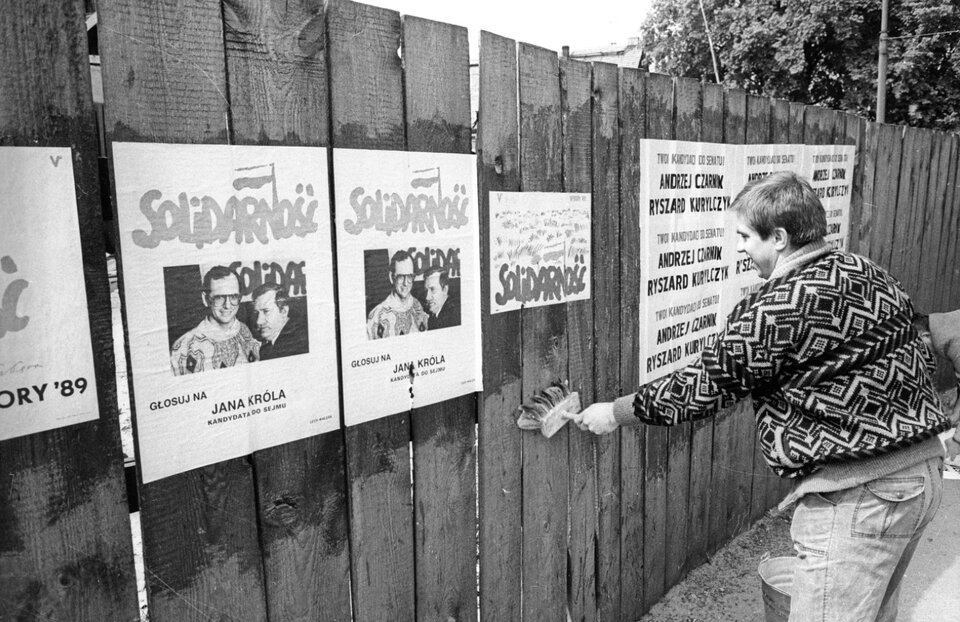 Zdjęcie przedstawia mężczyznę naklejającego na drewnianym płocie plakaty. Plakaty zachęcają do głosowania na Jana Króla należącego do Solidarności.   