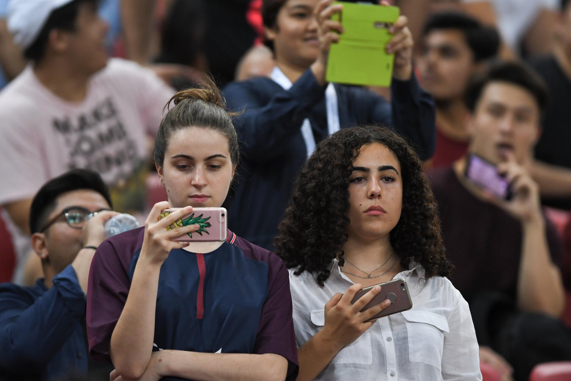 Zdjęcie przedstawia dwie młode kobiety. Jedna trzyma w dłoni telefon, patrzy na ekran, nagrywa mecz. Druga trzyma telefon i nagrywa, ale patrzy w stronę murawy.  Obie znajdują się na trybunach na stadionie. Obie mają poważne wyrazy twarzy. 