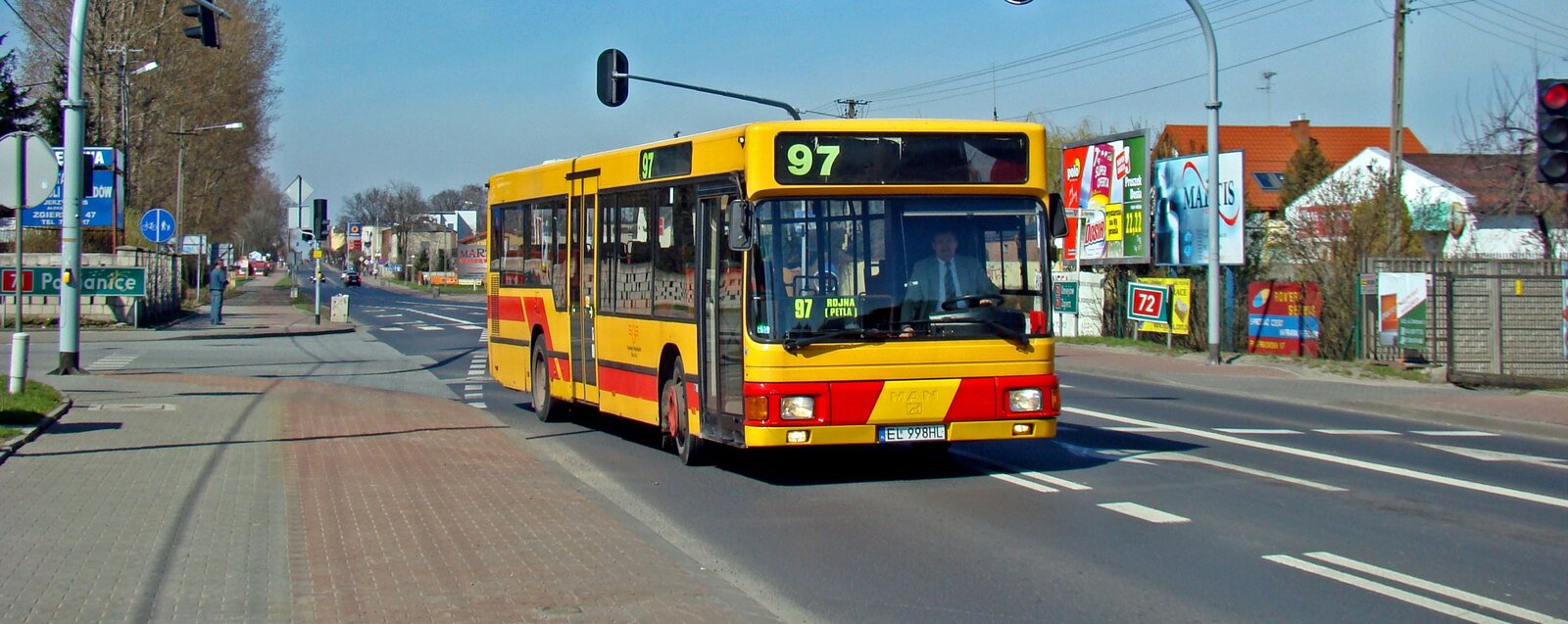 Zdjęcie przedstawia widok ulicy. Żółto-czerwony autobus o numerze 97 jedzie miejską ulicą. Wzdłuż jezdni stoją znaki drogowe, sygnalizatory świetlne oraz drzewa. Po prawej stronie drogi widać budynki mieszkalne, ogrodzenia, tablice z reklamami.