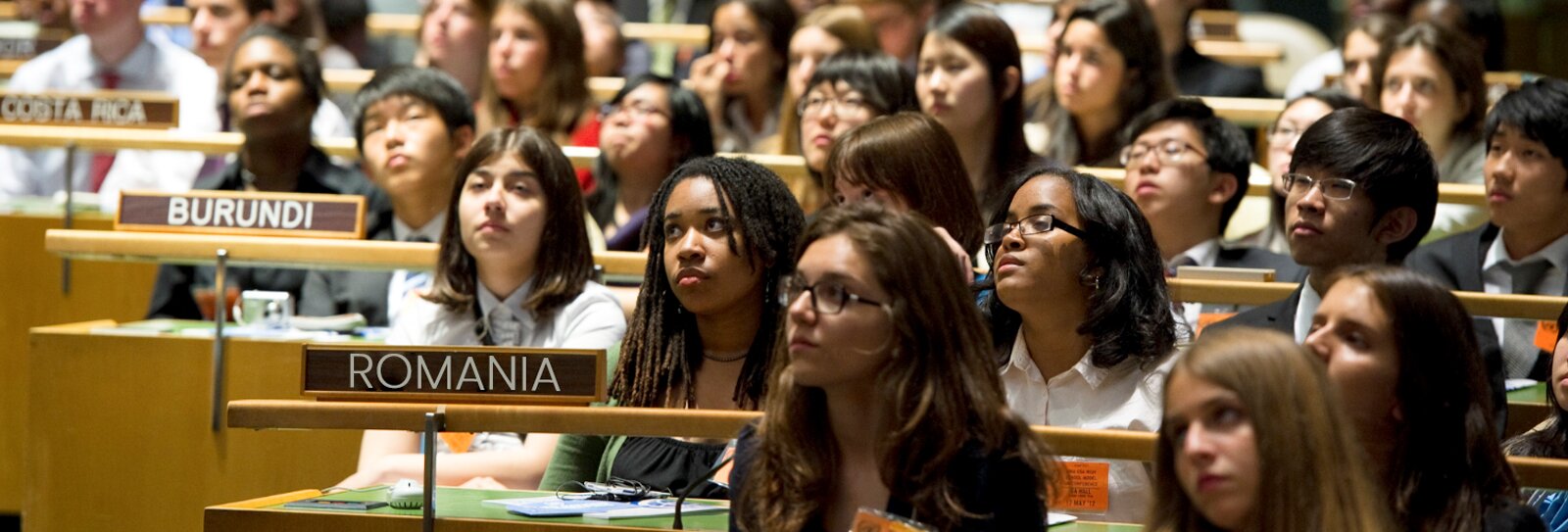 Zdjęcie przedstawia młode osoby siedzące w rzędach. Głowy wszystkich zwrócone są w tym samym kierunku. Każdy rząd ma tabliczkę z nazwą kraju jaki reprezentuje. Widoczna jest tabliczka z nazwą Romania, Burundi i Costa Rica. Transkrypcja znajduje się poniżej