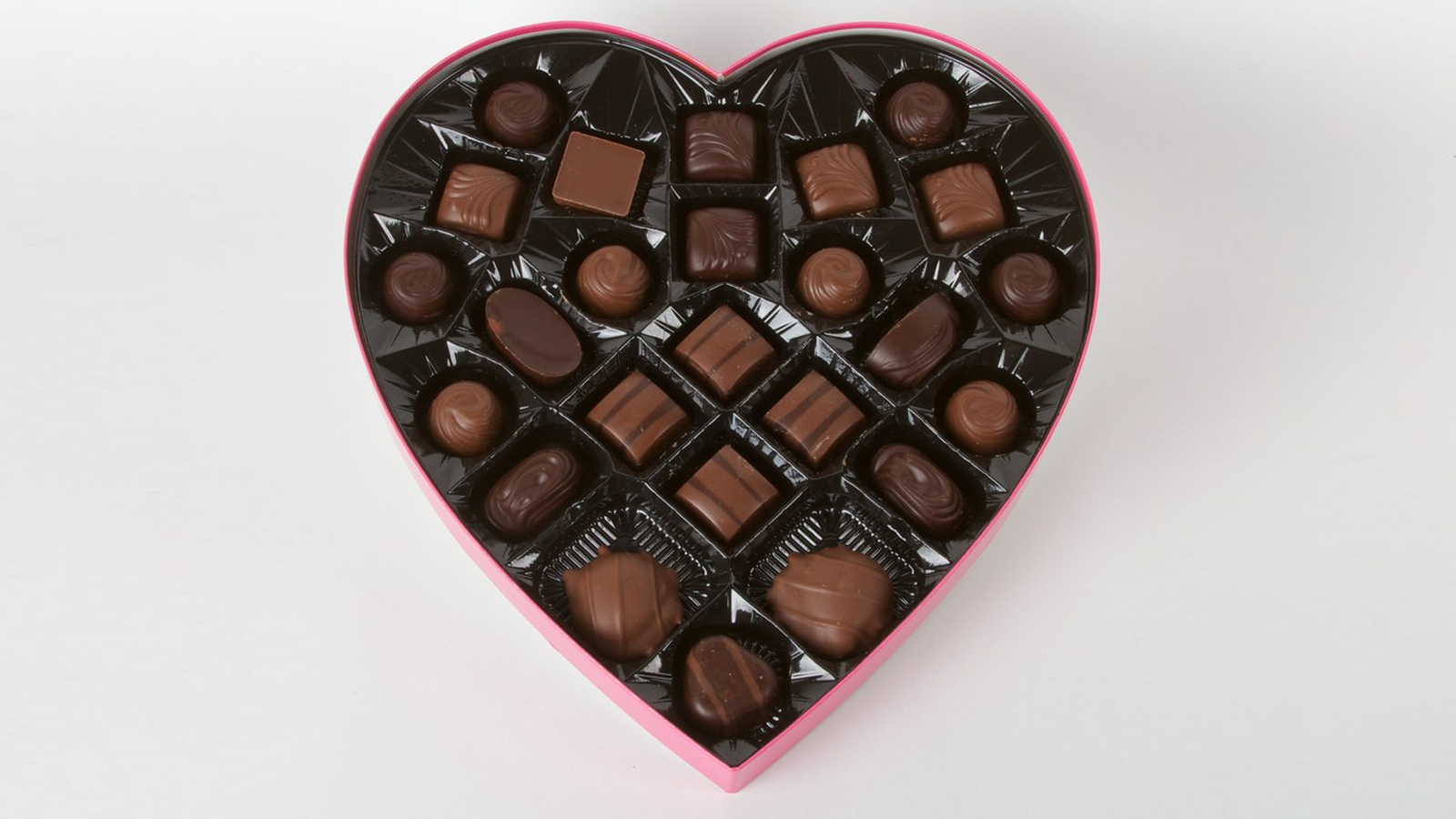 Zdjęcie przedstawia pudełko w kształcie serca wypełnione małymi czekoladkami w różnych kształtach, wykonanymi z ciemnej i mlecznej czekolady. 