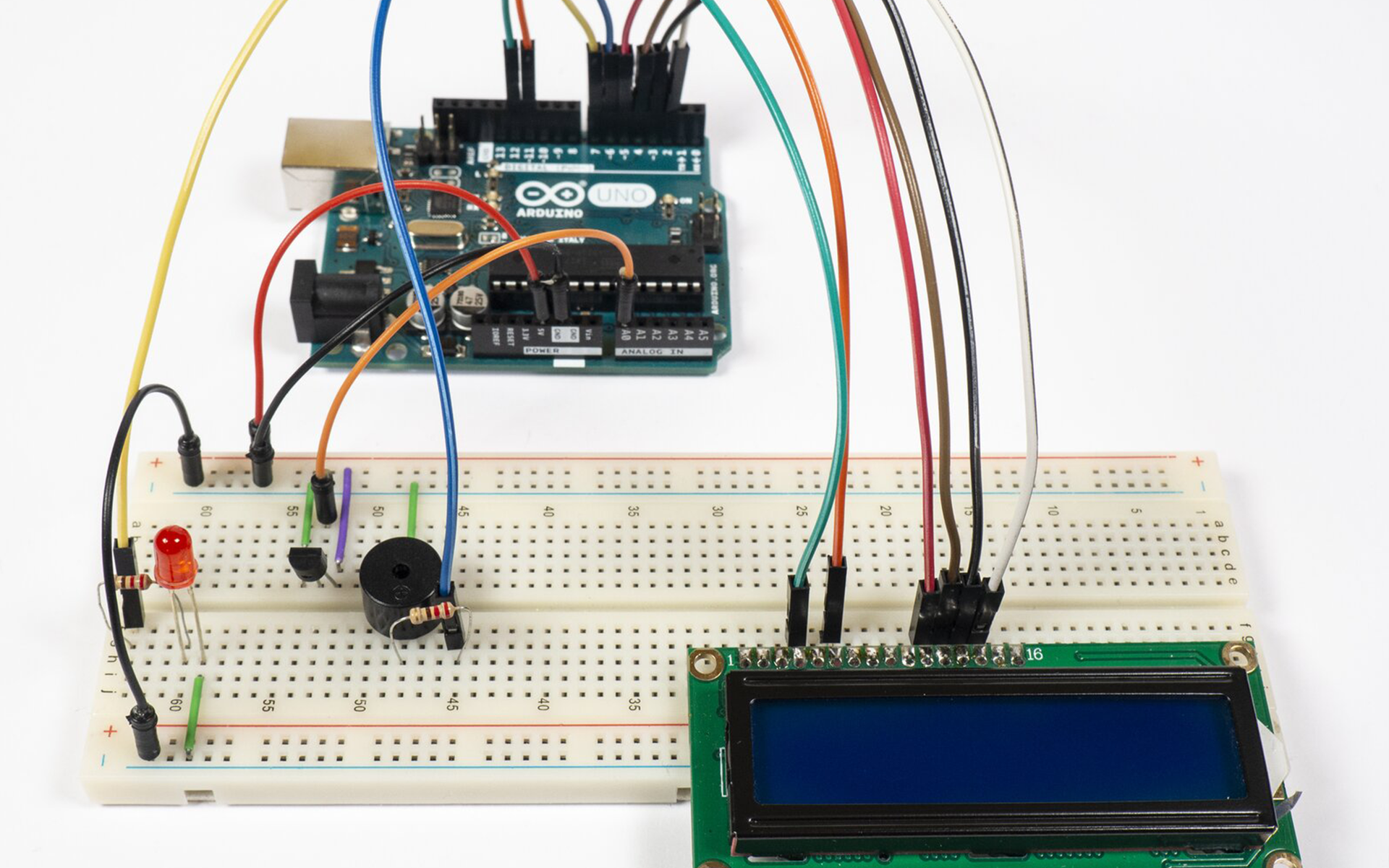 Zdjęcie przedstawia połączenia pomiędzy arduino uno, a płytką stykową. Na płytce znajduje się termometr (względem wycięcia w obudowie) podłączony jest prawą nóżką do uziemienia, lewą do +5V a środkową do pinu A0 arduino. Pin 5V podłączony jest do +5V płytki, a GND podłączone jest do uziemienia. Uziemienie górnej szyny jak i dolnej na płytce stykowej jest połączone. Podłączona jest także LED, jedną nóżką do uziemienia, drugą zaś poprzez rezystor do pinu 7 arduino. Po środku płytki zamontowany jest brzęczyk, który poprzez rezystor łączy się z pinem 6 arduino, a drugą nóżką do uziemienia. Po prawej stronie płytki stykowej podłączono ekran LCD, do którego podłączono kable prowadzące do arduino.