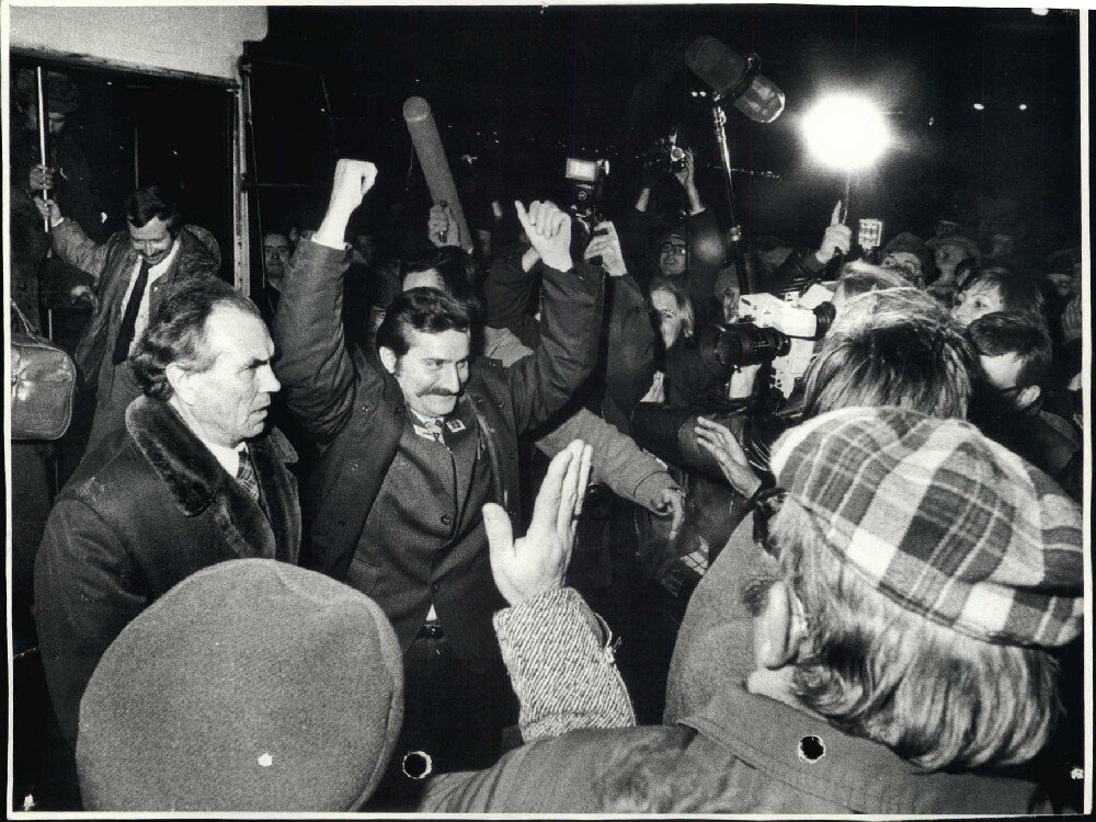 Ilustracja przedstawia cieszącego się Lecha Wałęsę, unoszącego ręce w zwycięskim geście. Obok niego znajduje się duży tłum. Zebrani są kamerzyści, dziennikarze. 