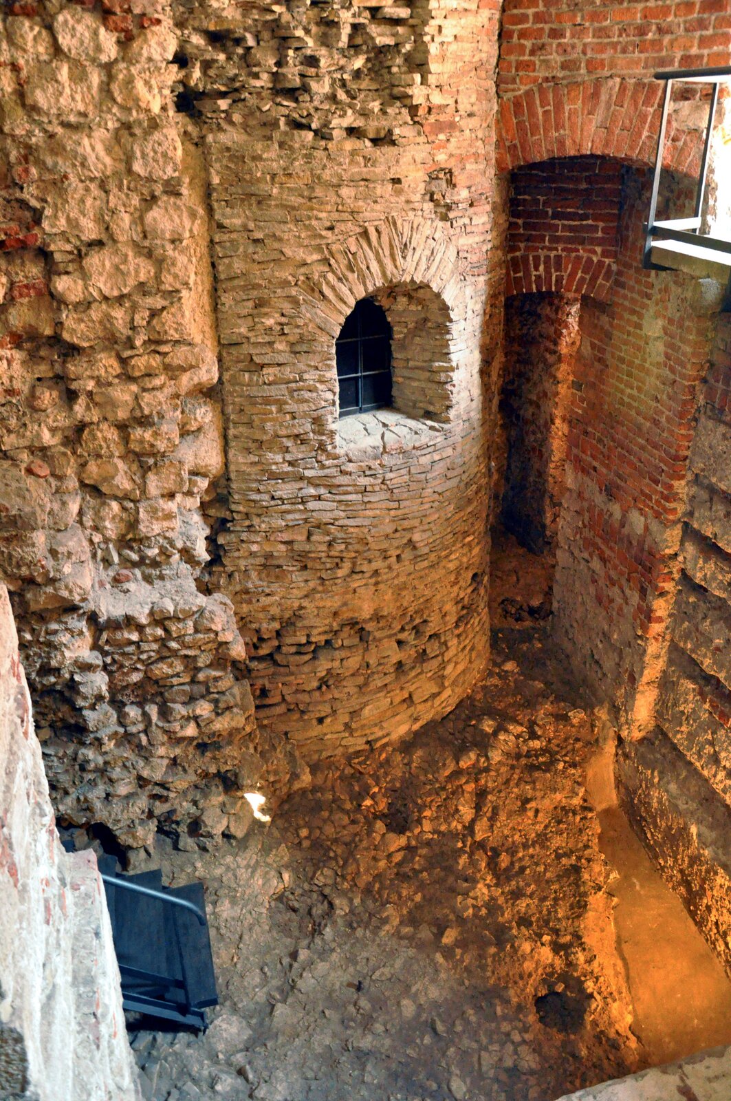 Ilustracja przedstawia fragment rotundy, wieży zbudowanej na planie koła, z niewielkim oknem z kratami o łukowatym zwieńczeniu. Rotunda przylega do murów dalszych zabudowań.