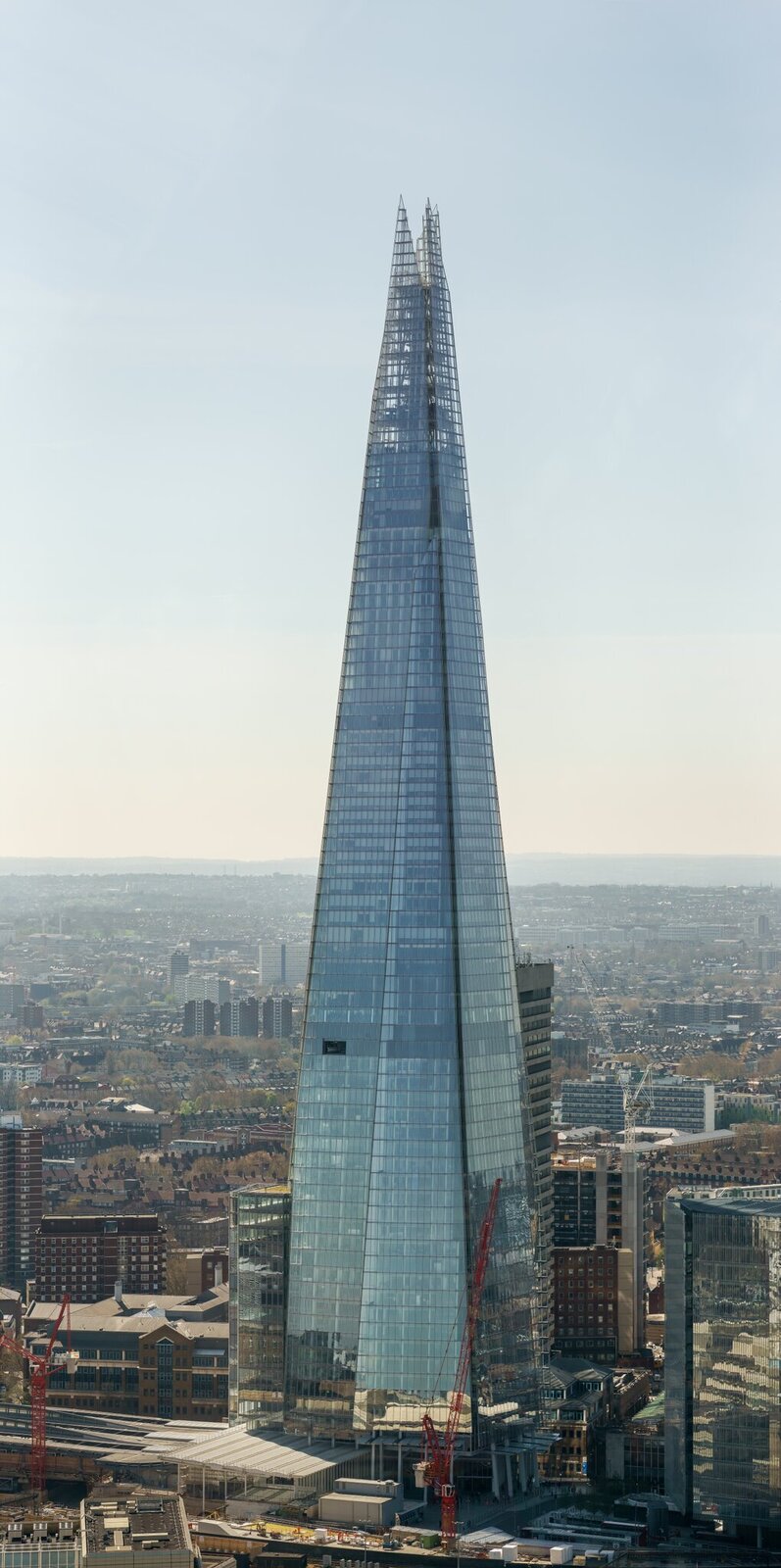 Ilustracja przedstawia wieżowiec The Shard na tle budynków Londynu oraz jasnego nieba. Góruje on nad widoczną w kadrze architekturą miasta, znacznie przewyższając pozostałe budynki. Od podstawy aż po wierzchołek, wieżowiec zbudowany jest ze szkła. Tworzą go szyby, które odbijając światło, zdają się być niebieskie. Ściany składają się z połączonych ze sobą wielu mniejszych, prostokątnych szyb. W dolnych partiach wieżowca można zauważyć lustrzane odbicia stojących przed nim jasnych budynków. Im wyżej, tym wieżowiec jest węższy, a jego wierzchołek dzieli się na dwie części, przypominające ostro zakończone trójkąty. 