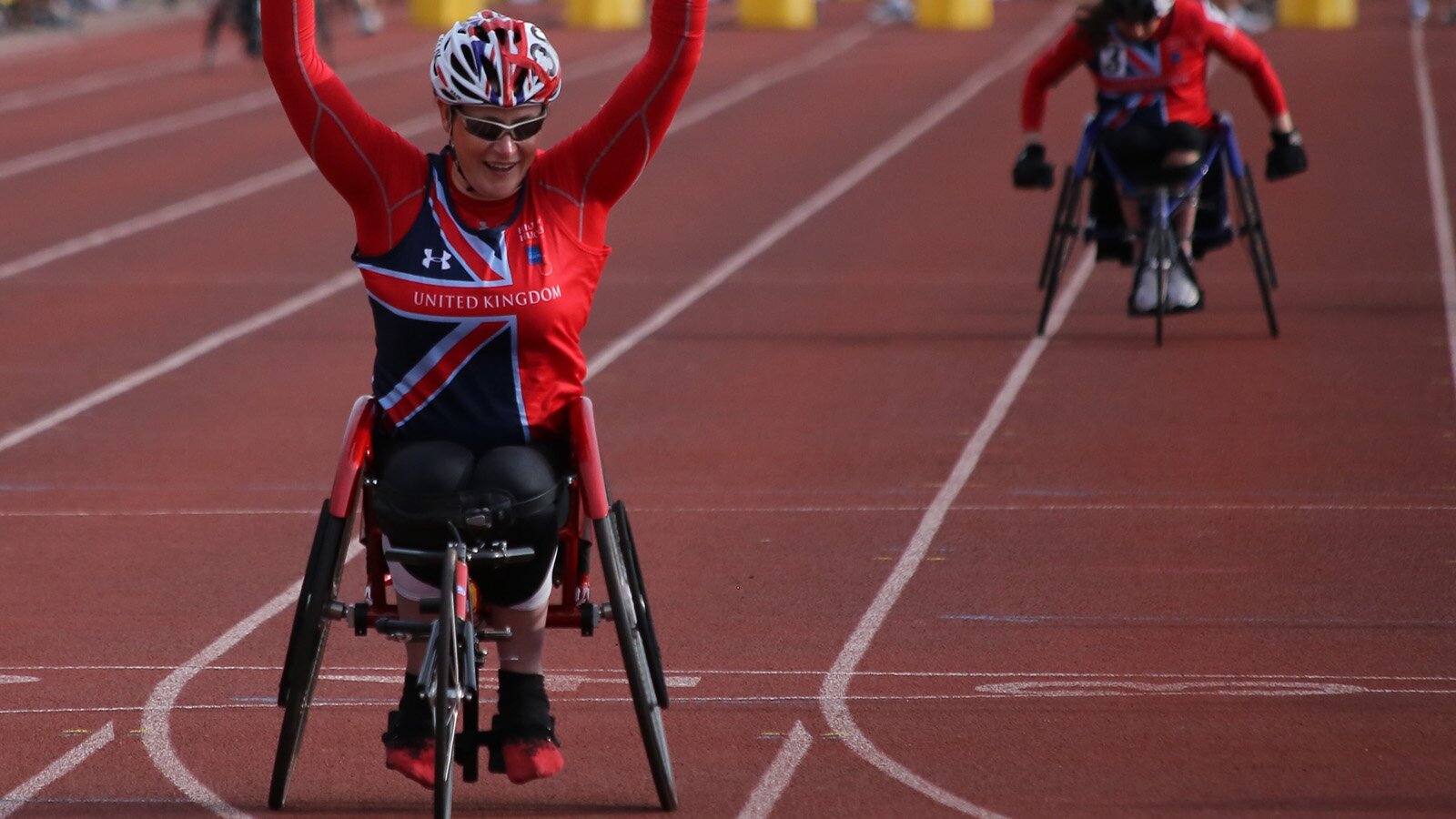Zdjęcie przedstawia dwóch zawodników na wózkach dla niepełnosprawnych, którzy biorą udział w zawodach odbywających się na bieżni. Osoby ubrane są w sportowe stroje: obcisłe bluzki, legginsy, kaski oraz sportowe obuwie.