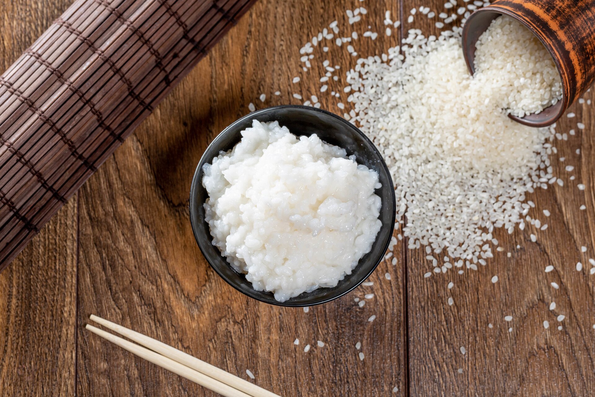 Zdjęcie przedstawia ryż. Ugotowany ryż znajduje się w miseczce. Natomiast nieugotowany wysypuje się obok z puszki na stół. Na stole leżą jeszcze bambusowe pałeczki i mata.