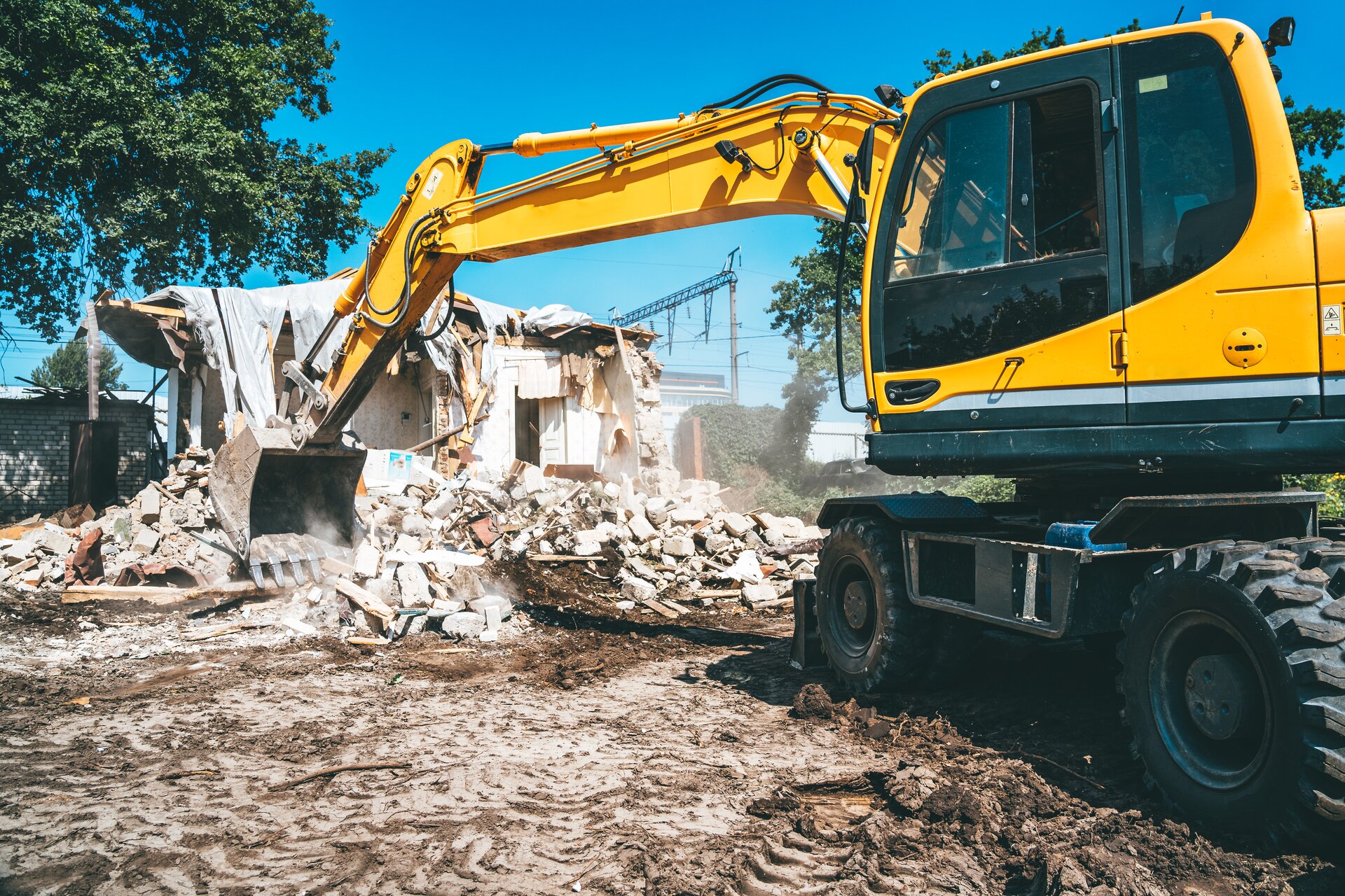 Zdjęcie przedstawia koparkę podsiębierną. Widoczna jest żółta koparka, w ujęciu z boku, stojąca na rozjeżdżonej ziemi. Ramię koparki jest wyprostowane, a łyżka nabiera gruz z rozwalonego budynku znajdującego się w tle. Po prawej stronie od ramienia widoczna jest kabina operatora koparki. W tle widoczny jest budynek podczas rozbiórki.

Opisy obiektów prezentowanych na grafikach są załączone w treści galerii zdjęć.