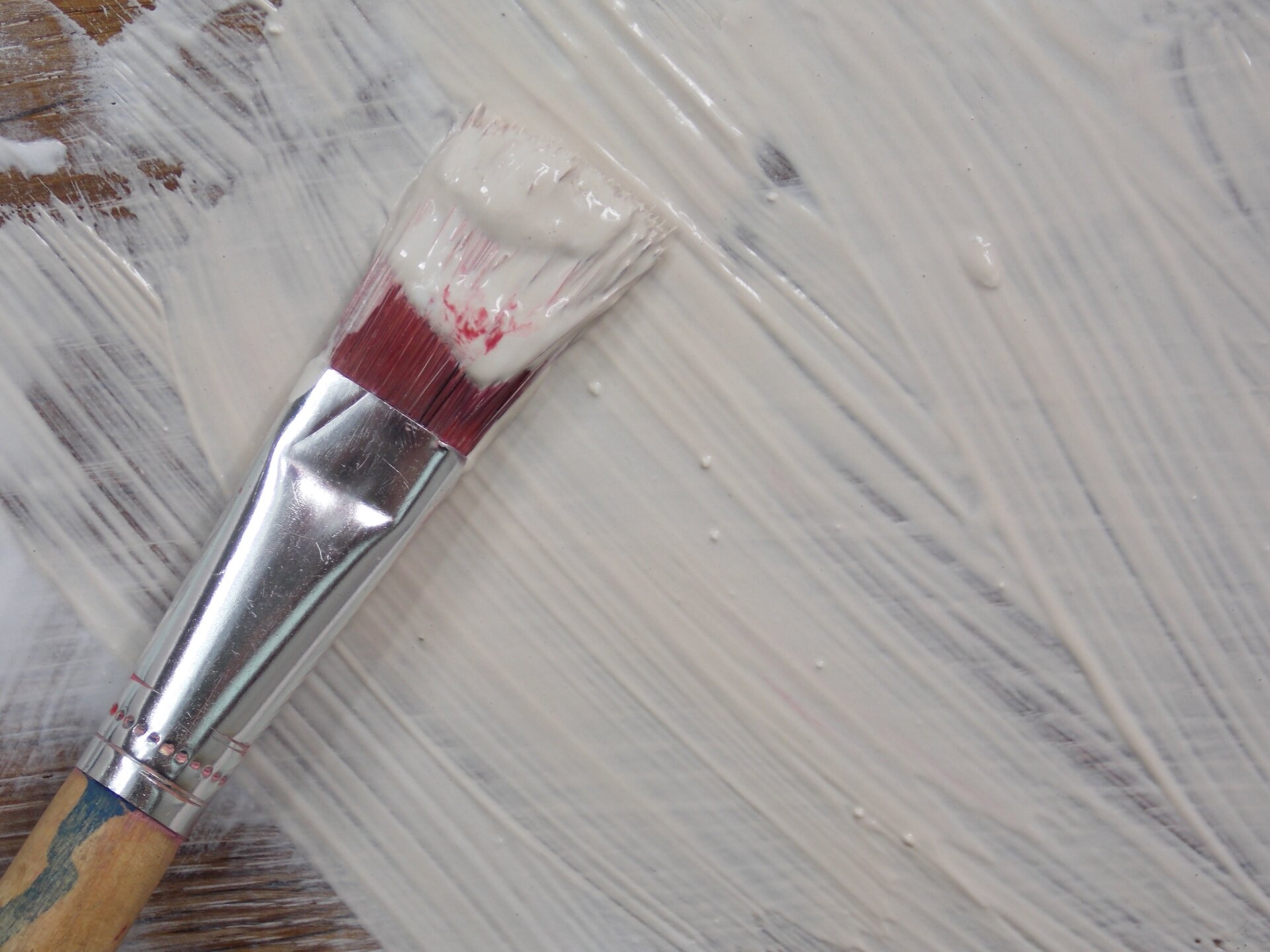 Ilustracja przedstawia pędzel z czerwonym włosiem, na którego końcu znajduje się farba. Tłem jest zamalowana na biało powierzchnia z widocznymi pociągnięciami pędzla.