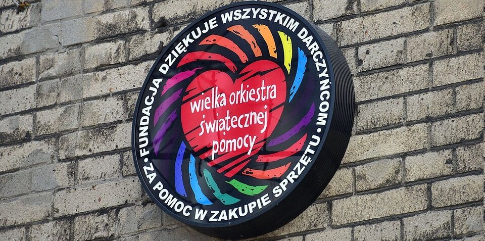 Ilustracja przedstawiająca logo WOŚP zawieszone na ścianie zewnętrznej budynku. Wkomponowane jest w czarny okręg z napisem: Fundacja dziękuje wszystkim darczyńcom za pomoc w zakupie sprzętu. Pośrodku znajduje się duże czerwone serce z napisem: wielka orkiestra świątecznej pomocy. Z serca rozchodzą się półkoliste linie w różnych kolorach.