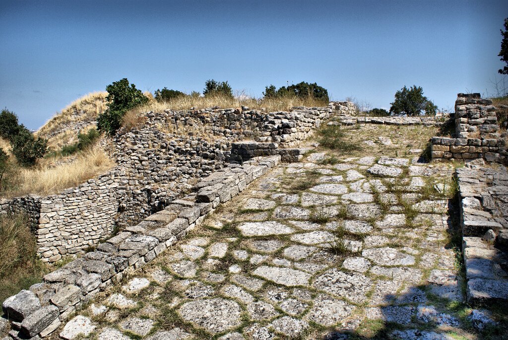 Zdjęcie kolorowe przedstawia kamienną konstrukcję delikatnie porośniętą częściowo wyschniętą trawą. Budowla wykonana jest z płaskiego kamienia, dobrze dopasowanego do siebie, który tworzy mury i chodniki o niezbyt szerokich ścieżkach, w tle małe drzewka.