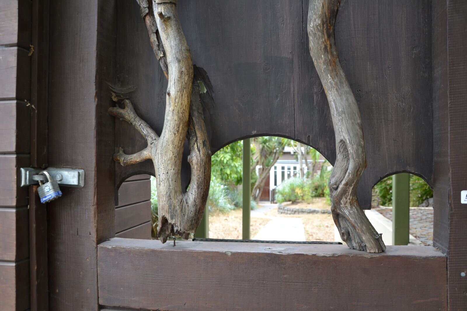 Ilustracja przedstawia fotografię furtki wejściowej do ogrodu. Są w niej otwory, przez które widoczne są fragmenty drzew oraz zabudowań. Ozdobą metalowej furtki są konary drzew ułożone pionowo wokół otworów. Są bez kory i stanowią wyraźny akcent zwracający uwagę - głównie przez kontrast koloru i naturalnego materiału. 