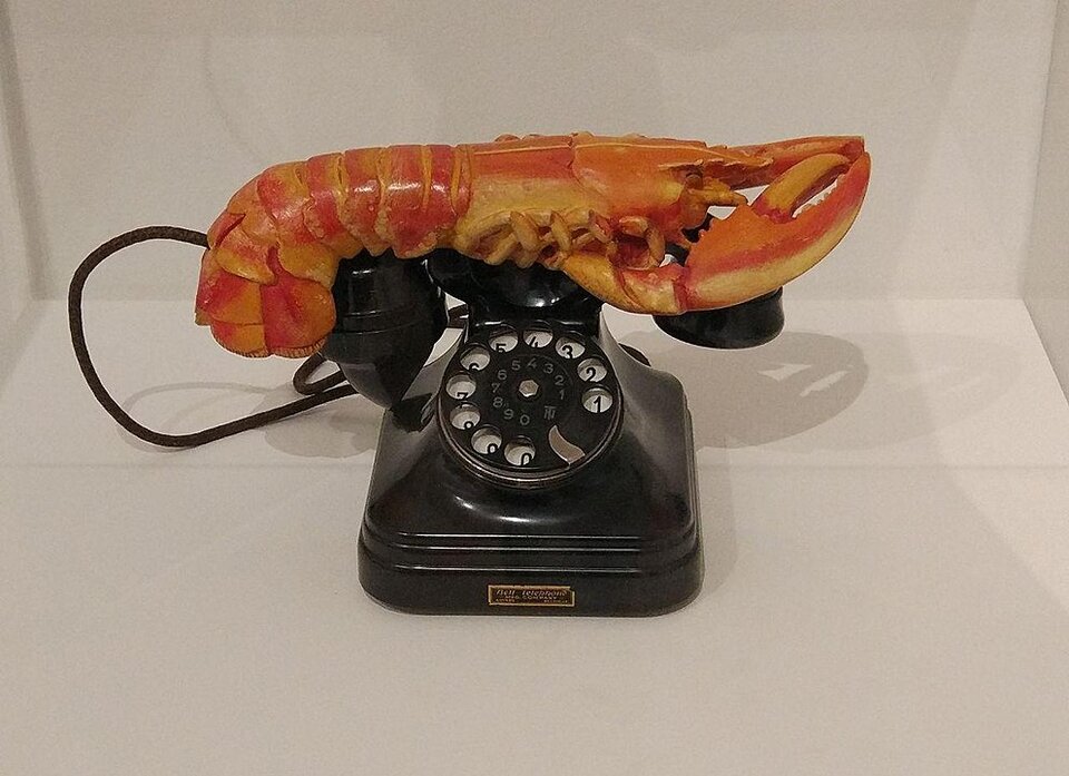 Zdjęcie przedstawia stojący na blacie aparat telefoniczny starego typu. Na wierzchu urządzenia znajduje się okrągła tarcza z dziesięcioma niewielkimi otworami. Są one oznaczone cyframi od 1 do 0. Za tarczą umieszczone są widełki, na których opiera się słuchawka. Rączka słuchawki jest ozdobiona rzeźbą wyobrażającą leżącego homara. Głowa i szczypce zwierzęcia znajdują się po stronie słuchawki, a ogon po stronie mikrofonu.