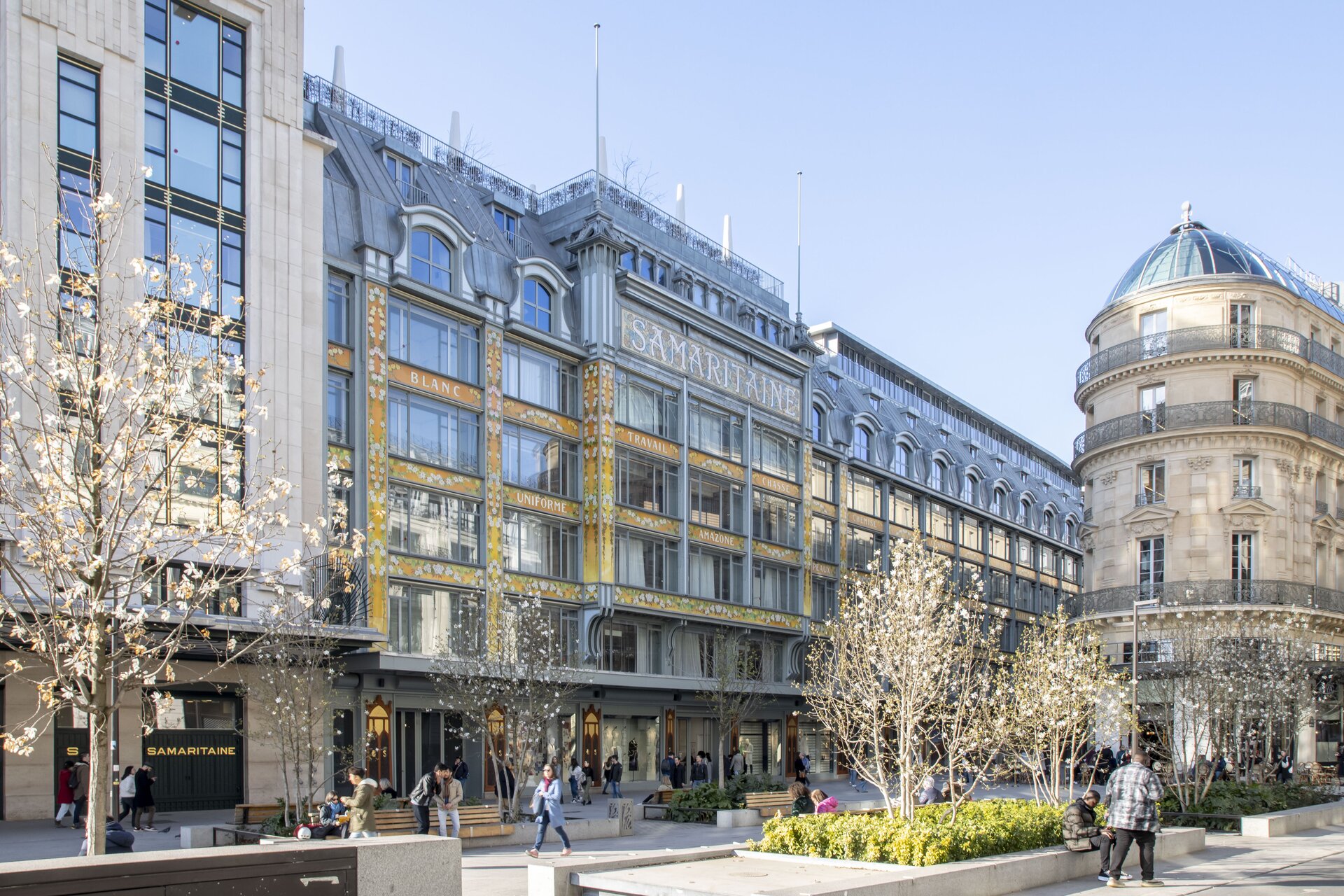 Ilustracja przedstawia centrum miasta. Kamienice i budynki handlowo usługowe oraz szeroki deptak.