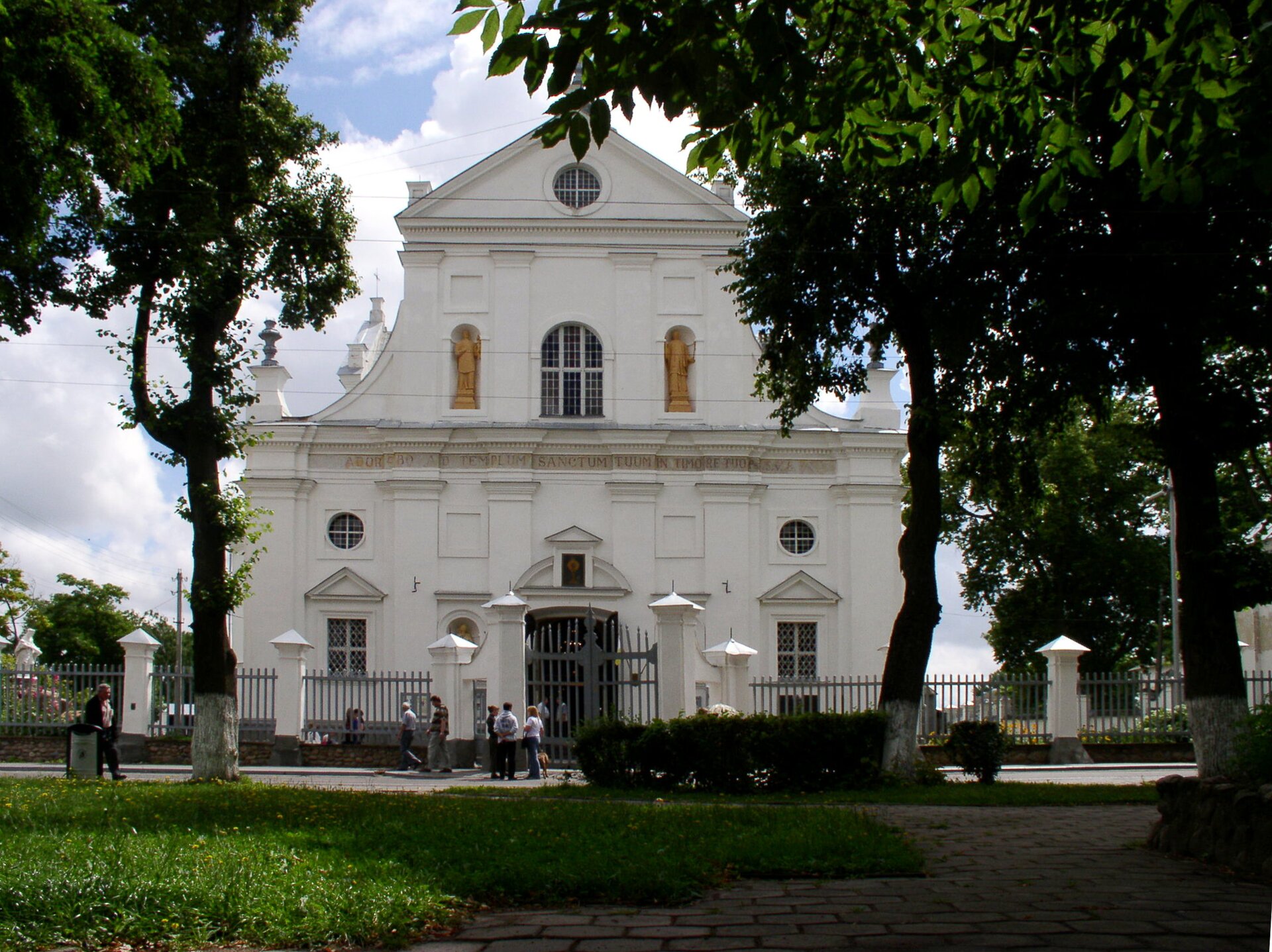 Zdjęcie przedstawia białą fasadę kościoła. W centralnej części wejście, a z obu jego stron małe okna. Fasada przypomina kształt trójkąta równoramiennego, a w jego górnej części znajdują się okrągłe okna oraz dwie, równo rozmieszczone, żółte rzeźby przedstawiające świętych. Przed kościołem ogrodzenie, a przed nim droga, po której idą ludzie. Wokół zielone drzewa, krzewy i trawa. Niebo błękitne, częściowo przysłonięte białymi chmurami.