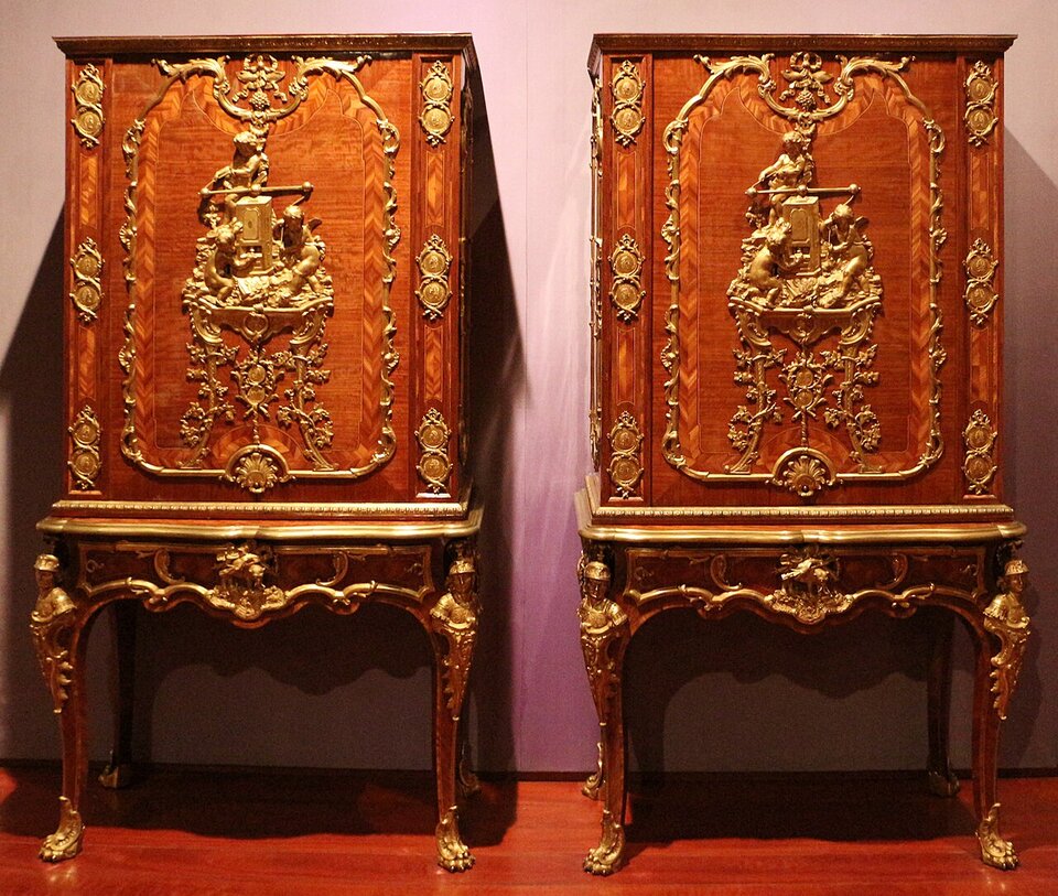 Ilustracja przedstawia dwie komody z drewna, stojące obok siebie, wykonane przez ebenistę Charles’a Cressenta. Są to komody na smukłych nogach, z przodu bogato rzeźbione finezyjnymi ornamentami, ze złoceniami. Środek każdej z nich zdobi płaskorzeźbiona scenka z postaciami, która podobnie jak pozostałe płaskorzeźbione ornamenty jest złocona. Komody są rokokowym meblem francuskim. który spopularyzował w Europie Charles Cressent w pierwszej połowie XVIII w.                