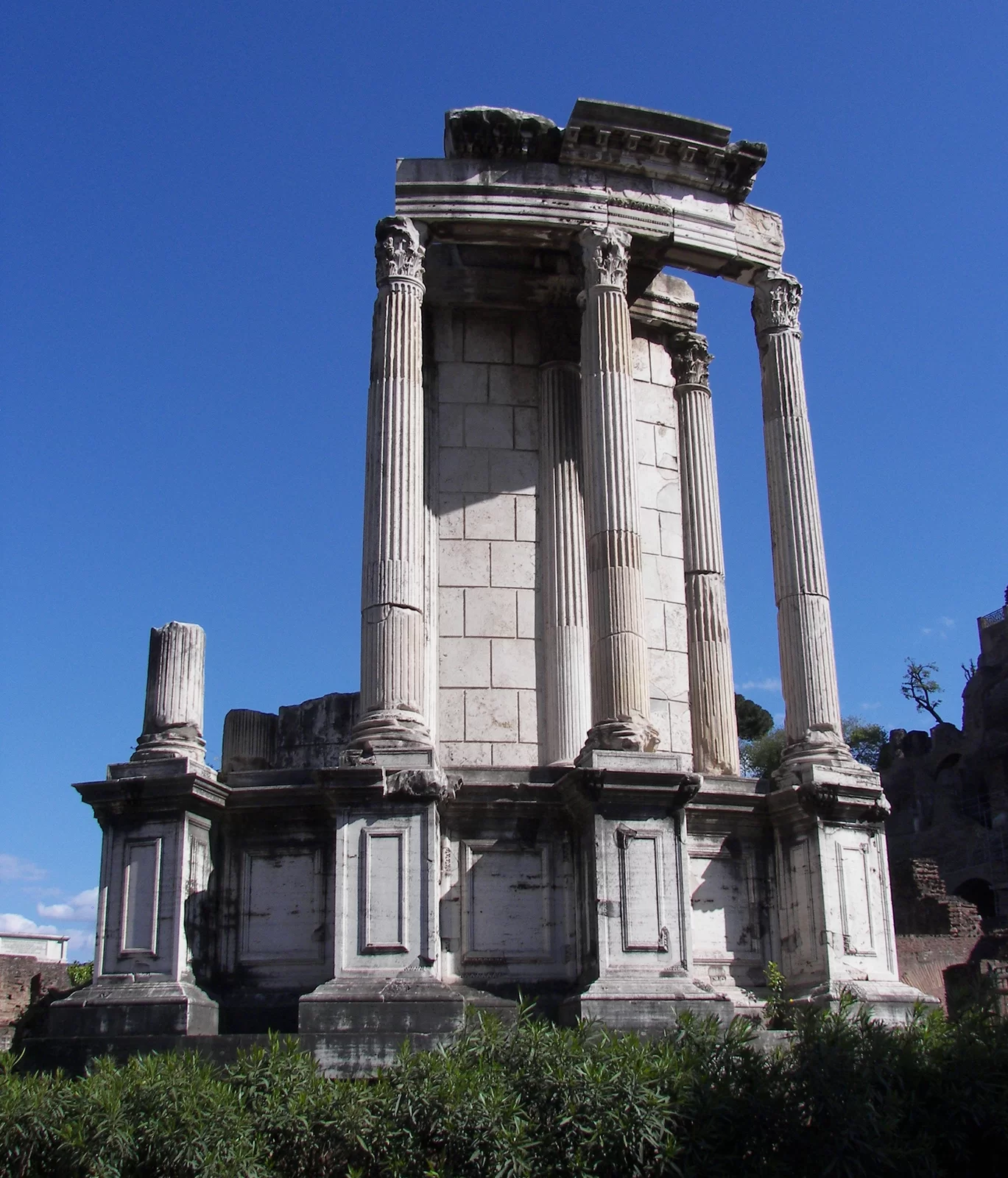 Fotografia przedstawia ruiny Świątyni Westy na Forum Romanum w Rzymie. Ukazuje trzy zachowane kolumny korynckie, które układają się w część pierścienia. Kolumny mają smukłe trzony i bogato zdobione kapitele korynckie z liśćmi akantu. Ustawione są na wysokim cokole. Na kolumnach znajduje się zachowany fragment belkowania.