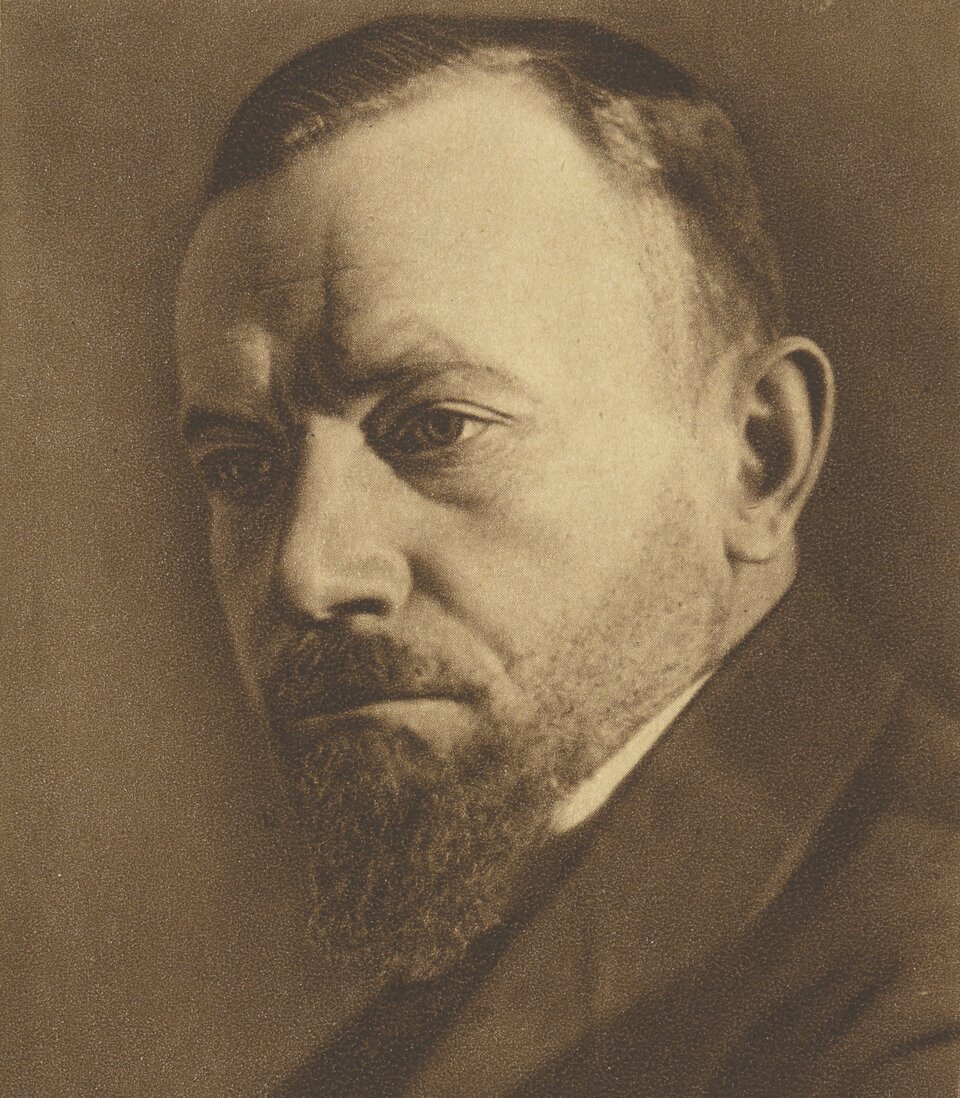 Zdjęcie przedstawia portret dojrzałego mężczyzny. Ukazany jest jego lewy profil. Mężczyzna ma pociągłą twarz z widocznymi zmarszczkami wokół oczu i na czole. Nos mężczyzny jest prosty, oczy wyraziste. Mężczyzna nosi krótko ostrzyżoną brodę i wąsy. Ubrany jest w garnitur i koszulę.