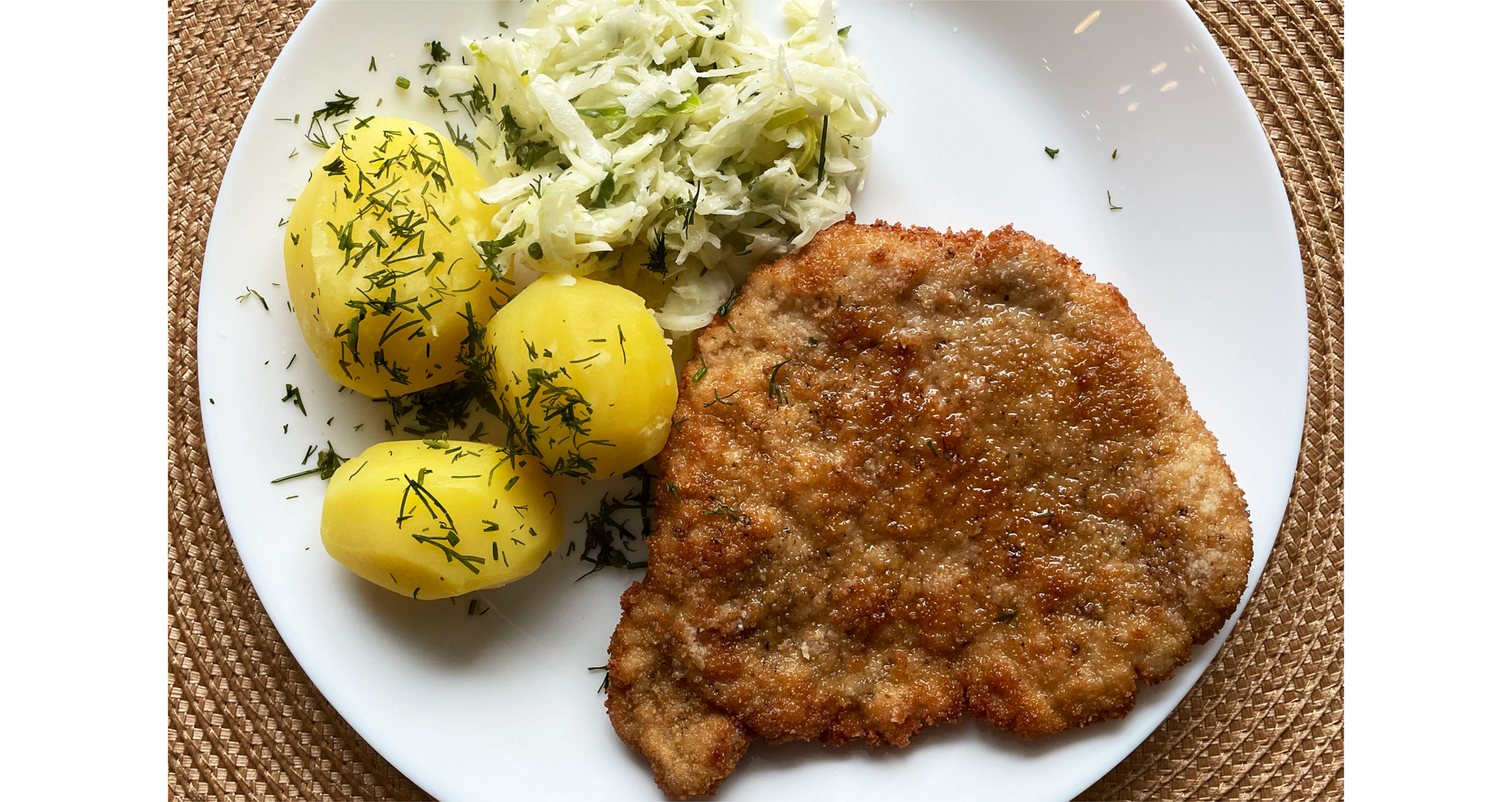 Zdjęcie przedstawia tradycyjne polskie danie na talerzu: schabowy, ziemniaki z koperkiem i surówkę z kapusty.