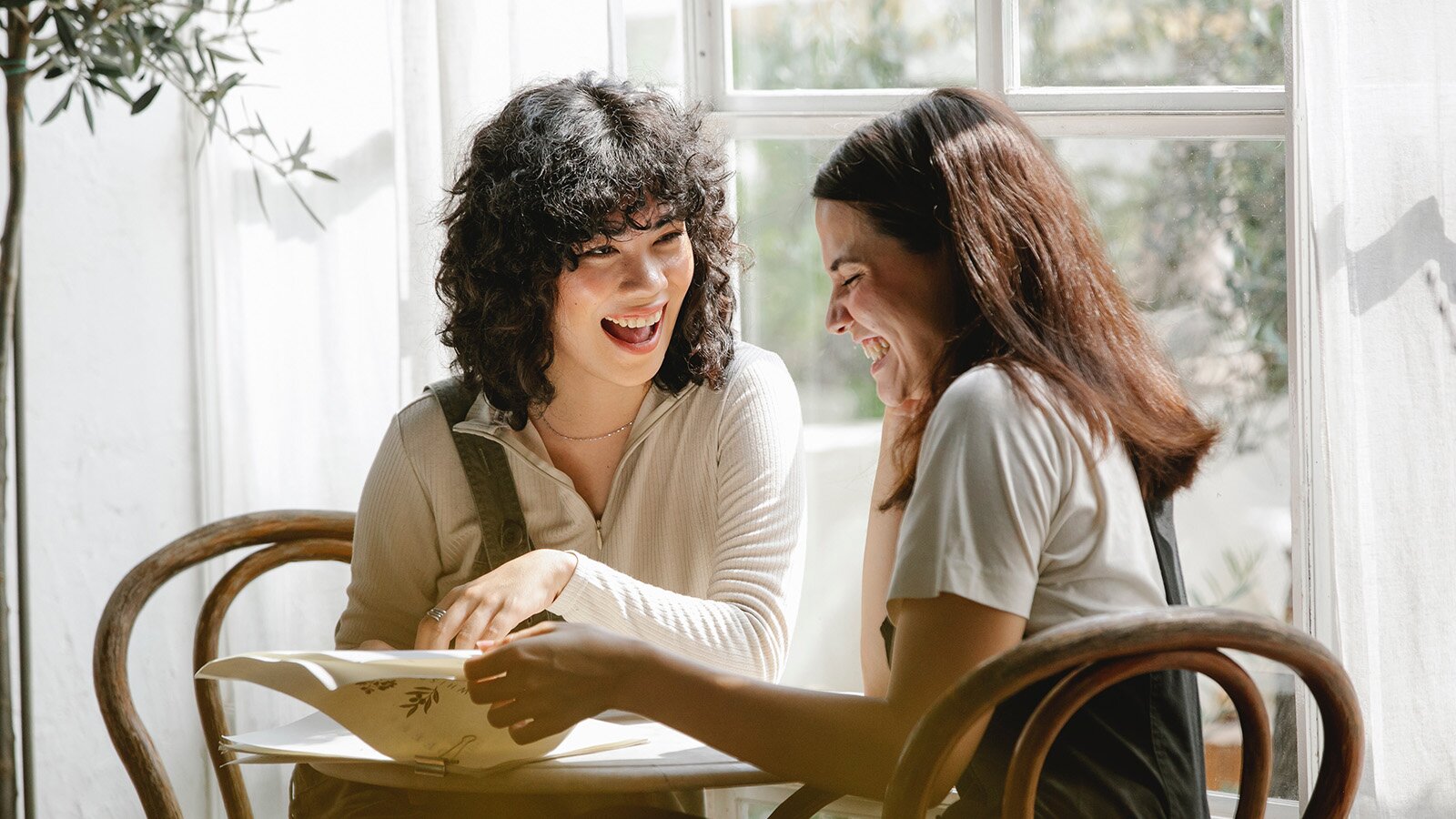 Zdjęcie przedstawia dwie kobiety w młodym wieku siedzące przy okrągłym stoliku, które przeglądają jakieś papiery. Obie uśmiechają się. 
