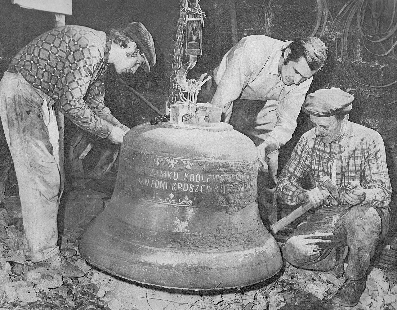 Fotografia przedstawia trzech mężczyzn pochylających się nad wielkich rozmiarów dzwonem. Mężczyzna po prawej stronie trzyma w ręku duży młotek. Dzwon zawieszony jest na haku. Widoczny jest na nim napis "Zamku królewskiego" i "Antoni Kruszewski".