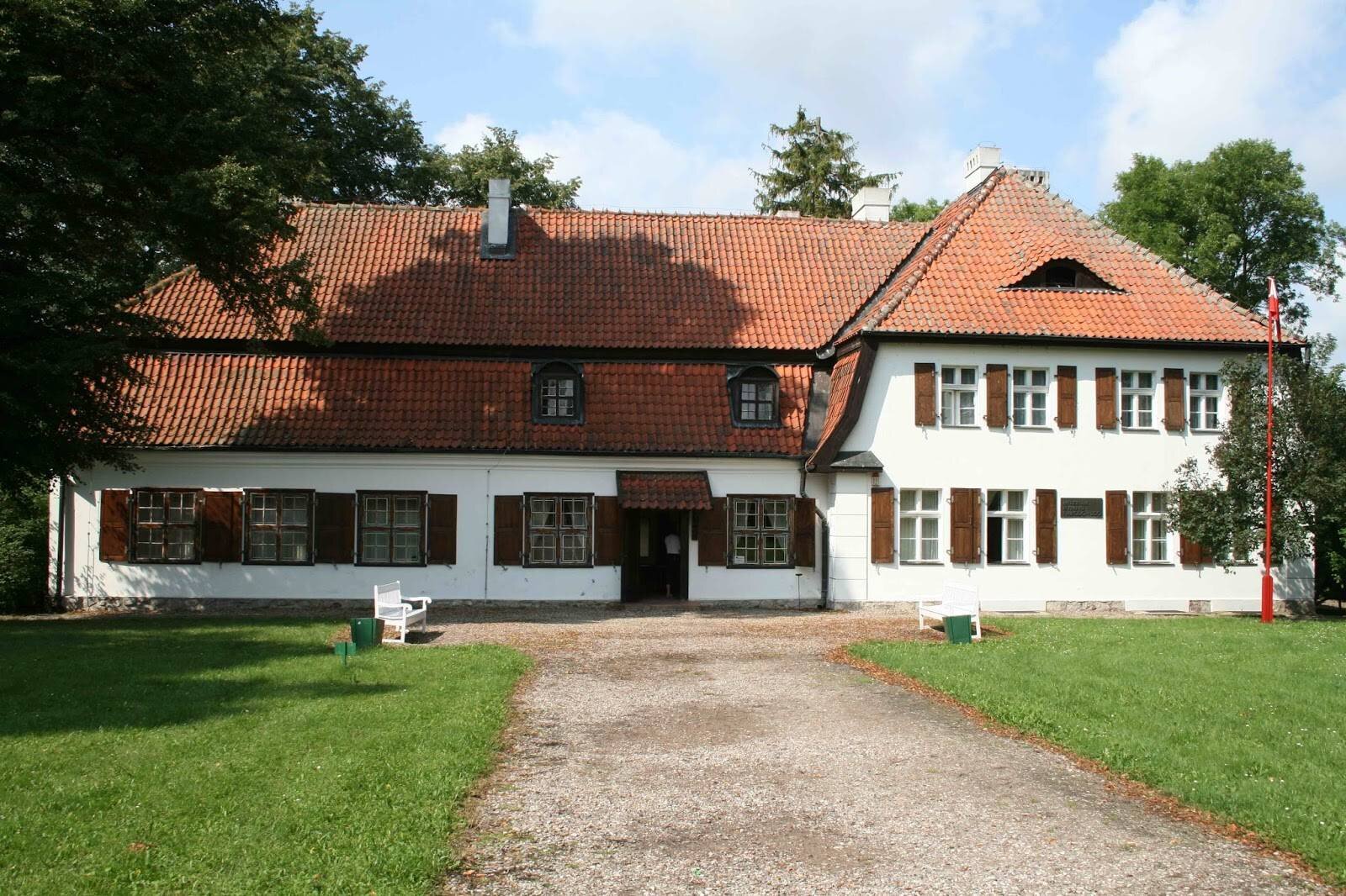 Na zdjęciu przedstawiony jest dwukondygnacyjny, rozległy budynek. Dach budynku jest spadzisty, pokryty dachówką. W centralnym punkcie ściany frontowej są drzwi, które przysłania niewielki daszek. Po lewej stronie budynku są okna kwadratowe z drewnianymi okiennicami. Po prawej stronie budynku są okna prostokątne z drewnianymi okiennicami. Dom otoczony jest drzewami. Prowadzi do niego szeroka żwirowa aleja. Przed domem stoją dwie ławki, z prawej strony znajduje się maszt z zawieszoną flagą.