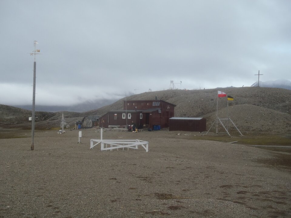 Zdjęcie przedstawia Polską Stację Polarną. Na pierwszym planie znajduje się fragment wydzielony niskim białym płotkiem oraz słup z kierunkami świata. Stacja składa się również z niewielkich brązowych zabudowań. Obok znajduje się maszt z flagami. Za budynkami znajduje się niewielkie wzniesienie, na którym stoi krzyż.
