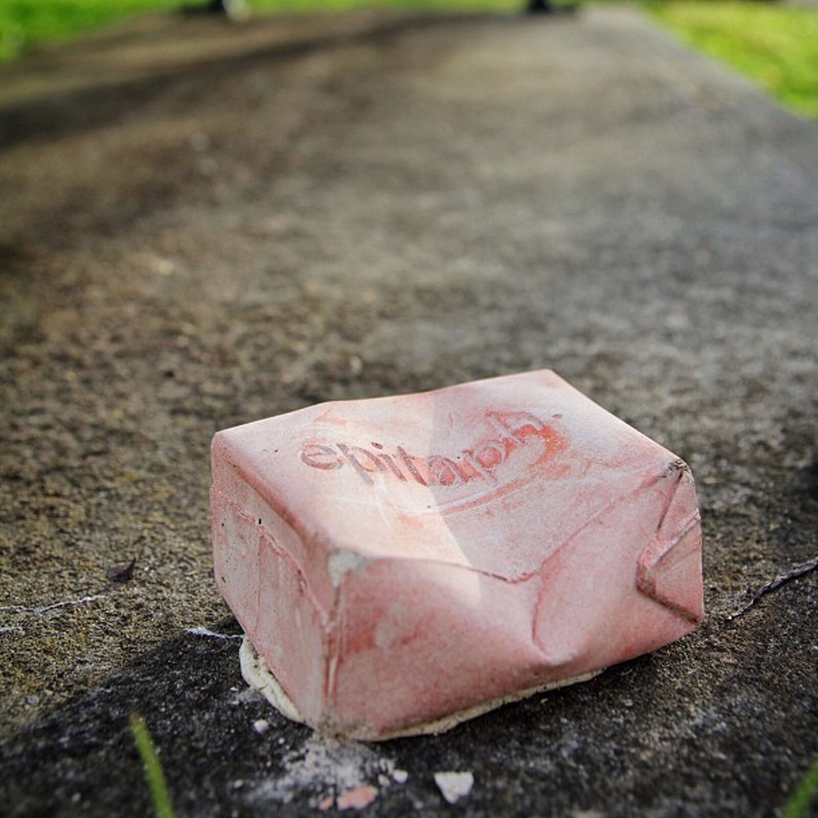 Ilustracja przedstawia prace Will Coles Epitaph. Ukazuje różowe, zgniecione pudełko, imitujące kartonowe opakowanie. Przedmiot leży na betonowym chodniku, w tle rośnie trawa. Na przedniej części kartonu znajduje się napis EPITAPH, co oznacza tablicę nagrobną "epitafium".