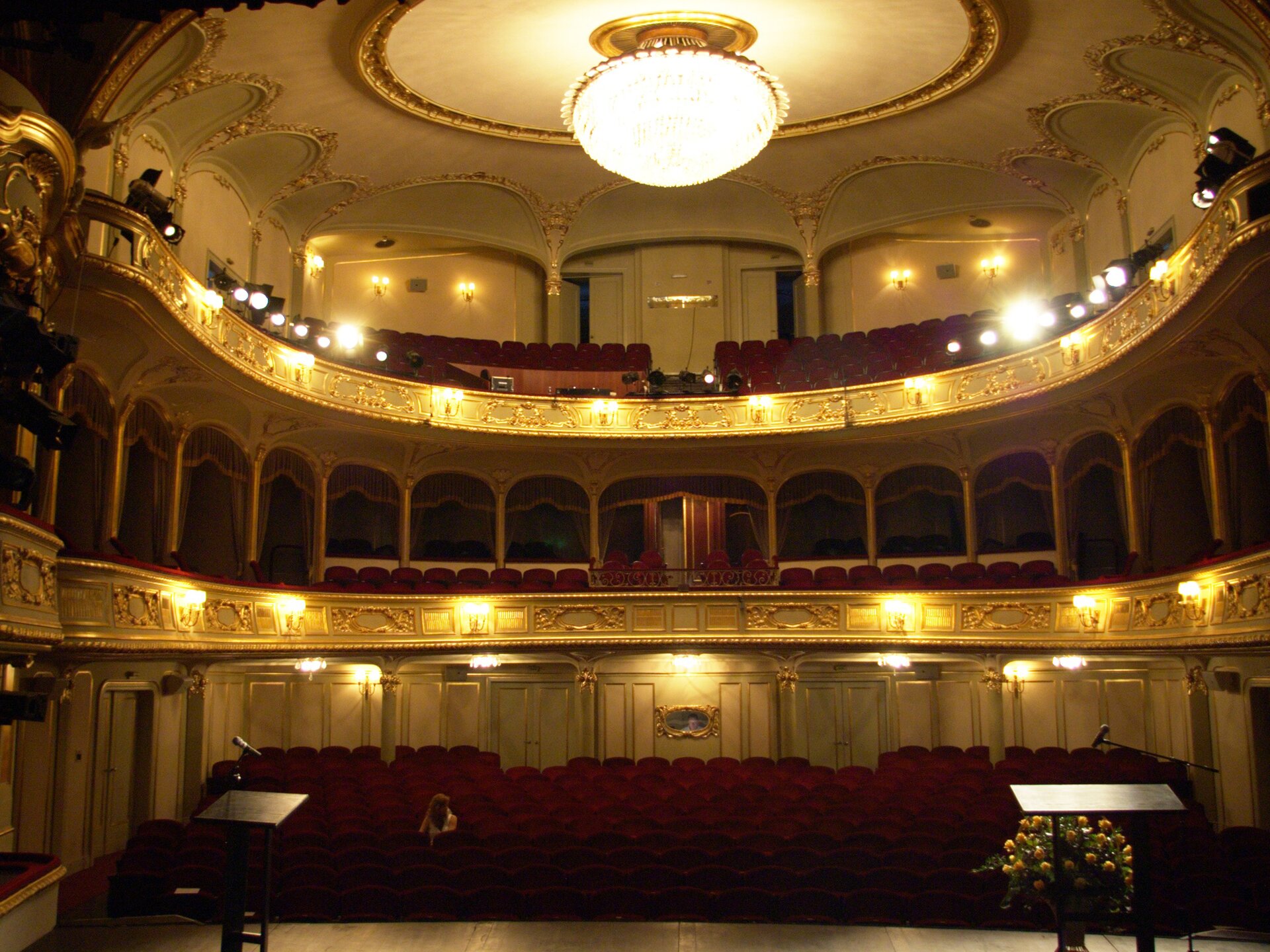 Fotografia barwna przedstawiająca widok ze sceny na widownię. Historyczne, pokryte białą farbą i złoconymi sztukateriami wnętrze teatru, posiada dwie kondygnacje wspartych na kolumnach balkonów. Ukształtowano je w podkowę, obejmującą również boczne ściany. Kolumn wspierających drugą kondygnację balkonów jest dwukrotnie więcej niż podtrzymujących pierwsze piętro z lożami, w tym większą, honorową, na środku. Po obu jej stronach, u dołu sąsiadujących z nią lóż, ustawiono w rzędzie po osiem foteli. Na drugim piętrze dwie kolumny wspierają strop nad widownią. Zdobiony złoconymi sztukateriami, ma on na środku duży okrągły plafon, otoczony złotą, rzeźbioną ramą. Rozświetla go ciepłym światłem kryształowy żyrandol, zwisający z również otoczonego złotą, lecz gładką ramą centralnego otworu plafonu. Ściany są otoczone łukami złoconej sztukaterii. Na bogato rzeźbionych balustradach balkonów jarzą się światła podwójnych kinkietów. Pięć podobnych lamp umieszczono na tylnej ścianie za fotelami na parterze, a cztery na ścianie wejściowej najwyższego balkonu. Na jego balustradzie po bokach ustawiono czarne reflektory. Ponieważ są włączone, na fotografii widać białe, okrągłe plamy świateł na ich zwróconych w stronę sceny na dole obiektywach. Loże balkonu pierwszego piętra zaopatrzono w ciężkie, bordowe zasłony. Tej samej barwy są wszystkie fotele w teatrze. Na lewo od środka ginącej w półmroku grupy foteli parteru bieleje samotna postać kobiety zasiadającej w drugim rzędzie widowni. Na pierwszym planie po bokach jasne prostokąty dwu pulpitów każdy z mikrofonem na statywie ustawianym na scenie, której skraj, wzdłuż dolnej krawędzi kadru, jasnym pasem odcina się na tle ciemniejszej widowni w tle. Za pulpitem z prawej strony, widoczny w pełni od strony widowni, duży bukiet żółtych róż w metalowym wazonie.