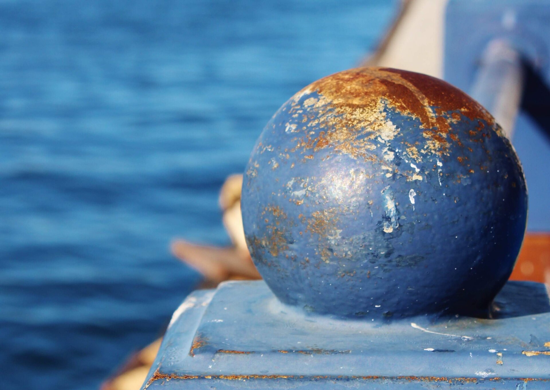 Zdjęcie przedstawia metalową kulę z odpryskami niebieskiej farby na powierzchni. Kula jest częścią poręczy pomalowanej na niebiesko, a w tle widać wodę, prawdopodobnie w jakimś porcie.