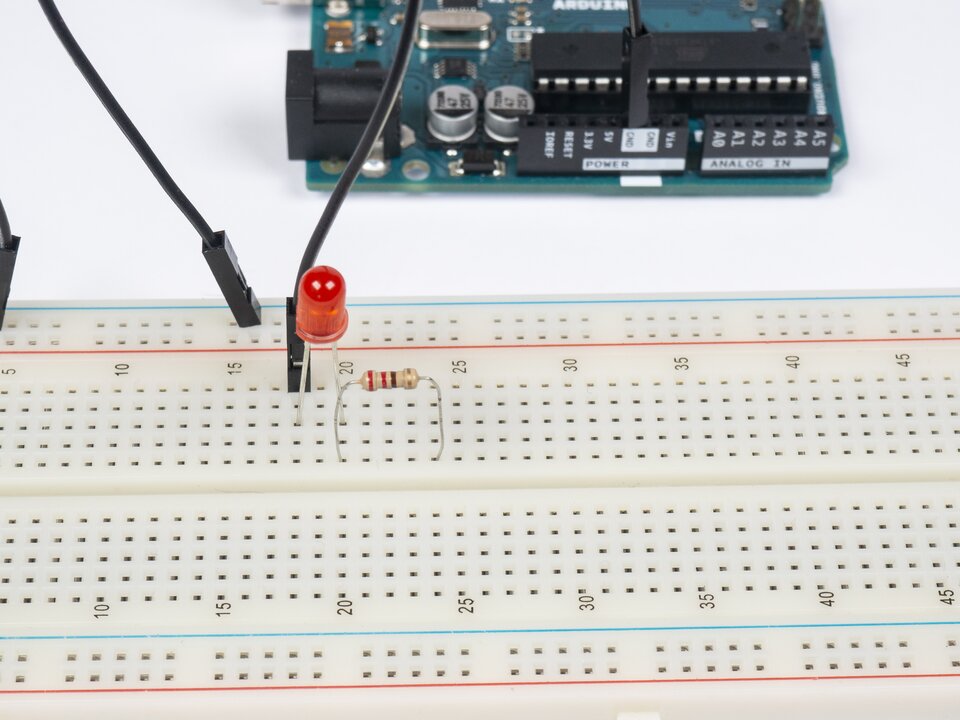 Zdjęcie przedstawia fragment płytki Arduino oraz fragment płytki stykowej. Wiersze podpisano co pięć. Na płytce stykowej znajduje się czarny przewód, czerwona dioda LED oraz rezystor. Rezystor ma na sobie linie w kolorach kolejno: czerwony, czerwony, brązowy, złoty. Ze złącza GND Arduino poprowadzono czarny przewód, którego drugi koniec umieszczono na szynie minus Arduino. Jedną nogę diody LED umieszczono w trzecim otworze wiersza 18, a drugą nogę w trzecim otworze wiersza 20. Jedną nogę rezystora, od czerwonego paska, umieszczono w piątym otworze wiersza 20, a drugą nogę umieszczono w piątym otworze wiersza 24.