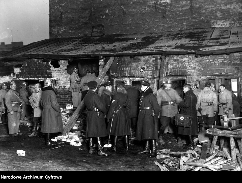 Zdjęcie przedstawia niewielki, parterowy budynek z cegły przylegający do innego, dużego budynku. Mały budynek jest częściowo zniszczony – ukazane są osmolone i pokruszone ściany oraz zniszczony dach. Przy budynku stoi duża grupa policjantów i strażaków. Mężczyźni oglądają budynek, rozmawiają ze sobą.