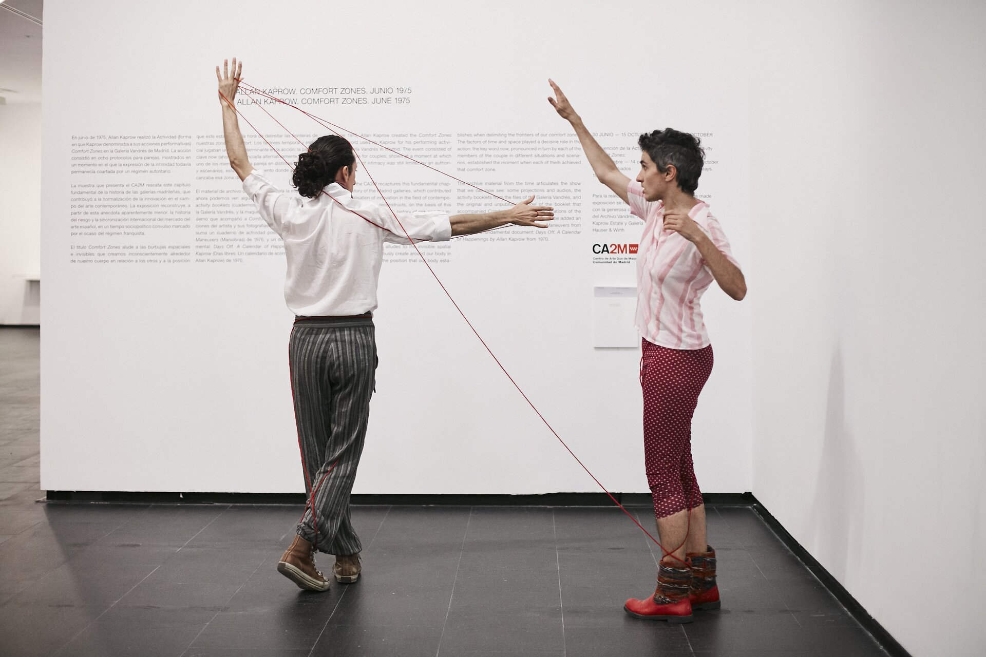 Zdjęcie przedstawia kobietę i mężczyznę w trakcie wykonywania performance'u. Mężczyzna stoi po lewej z uniesionymi rękami, ciągnie nimi za czerwonym sznurek. Sznurek jest owinięty wokół nogi stojącej po prawej kobiety, która ma uniesioną prawą rękę. Mężczyzna ubrany jest w białą koszulę i szare spodnie w paski, ma czarne włosy spięte w kitę. Kobieta ma krótkie włosy, czerwone spodnie i białą koszulkę w jasnoczerwone paski. Za postaciami stoi biała ścianka z napisami.