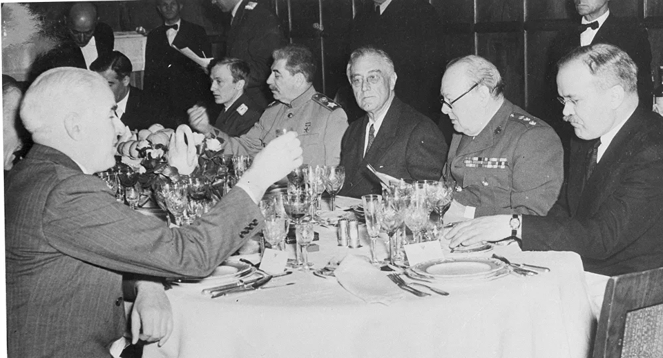 Zdjęcie przedstawia ośmiu mężczyzn siedzących wokół suto zastawionego stołu. Dwóch z nich podnosi do góry kieliszki wnosząc toast. Jednym jest krępy mężczyzna w średnim wieku, ubrany w mundur wojsk rosyjskich. Ma krótkie włosy, krzaczaste brwi i wąsy. Widzimy jego lewy profil. Drugim, siedzący po przeciwległej stronie, łysiejący mężczyzna ubrany w garnitur. Pozostali towarzysze obserwują rozwój wydarzeń. Jednym z nich, drugim z prawej, jest krępy, łysiejący mężczyzna w okularach, ubrany w mundur wojsk brytyjskich. Na naramiennikach kurtki ma naszyte trzy gwiazdki , a powyżej lewej kieszeni baretki. Obok niego po lewej stronie siedzi szczupły, starszy mężczyzna w okularach, ubrany w garnitur, a po lewej łysiejący mężczyzna w średnim wieku, o okrągłej twarzy. Ma on poważny wyraz twarzy, oczy lekko spuszczone w dół, zza cienkich okrągłych okularów i wąsy. Za biesiadnikami stoi pięciu mężczyzn, w tym obsługujący ich kelnerzy.