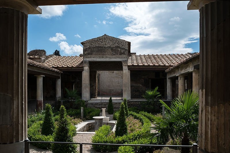 Ilustracja przedstawia wnętrze willi rzymskiej. Jest to kamienny budynek zbudowany na kształcie kwadratu z ogrodem pośrodku. Okna prowadzą na dzieciniec. Na dziedzińcu widać szereg niskich drzew, krzewów, drobnych roślin. Po bokach ogrodu rosną palmy. Na środku ogrodu znajduje się fontanna. Fontanna pozbawiona jest obecnie wody. Z czterech stron budynku znajduje się dziedziniec z kolumnami, który łączy się z ogrodem. Nad budynkiem znajduje się niebo z kłębiastymi chmurami.