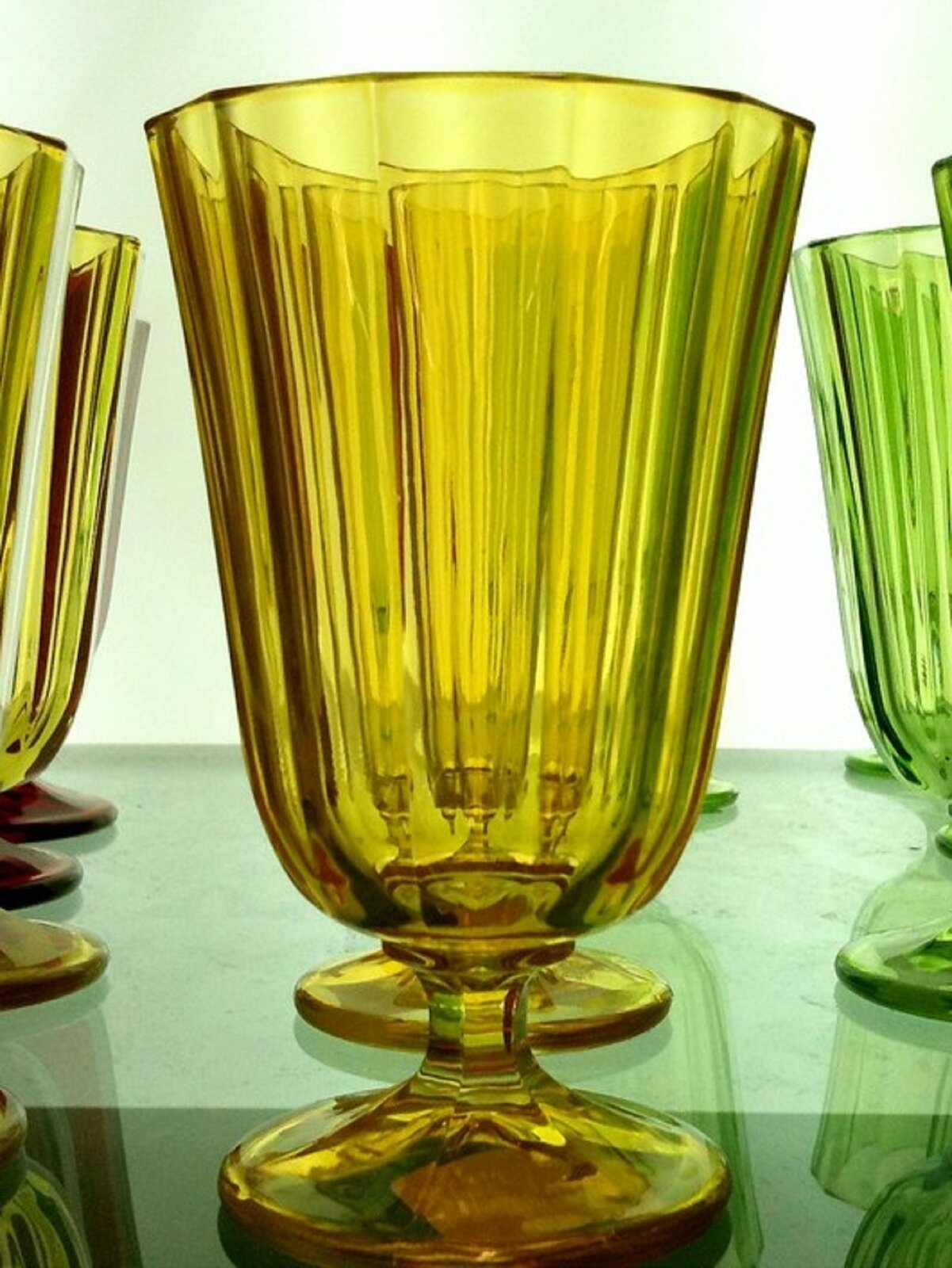 Grafika przedstawia szklane naczynie zabarwione na kolor żółty, stojące na rozszerzającej się ku dole nóżce. Posiada ono na swojej powierzchni podłużne żłobienia, a kształtem przypomina kielich.